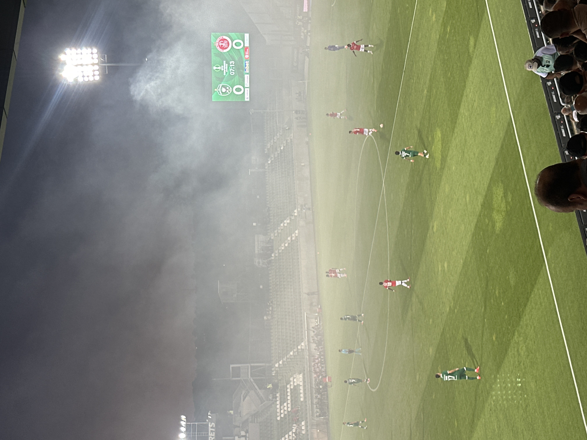 A football match is underway at Ludogorets Arena in Razgrad, Bulgaria, during a hazy evening. The stadium lights illuminate the pitch, where players in green and red kits are engaged in the game. The scoreboard displays "07:13" and a score of "0-0" between two teams, indicated by their respective emblems. Spectators are visible in the stands and a crowded area in the foreground. The atmosphere is somewhat obscured by smoke or fog, which is particularly dense around the stadium lights and the scoreboard.