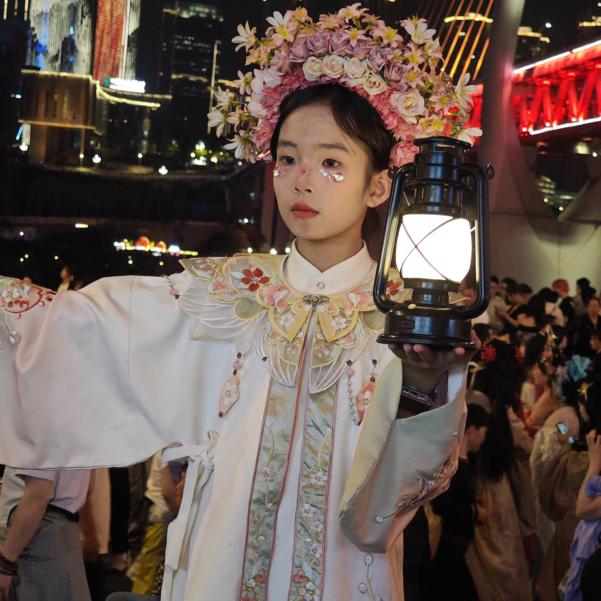 A young person stands holding a lantern in Yuzhong District, China. The individual is wearing an ornate white and gold robe, and a flower crown, with glitter around their eyes. Buildings are visible in the background. Many other people are standing behind the person.