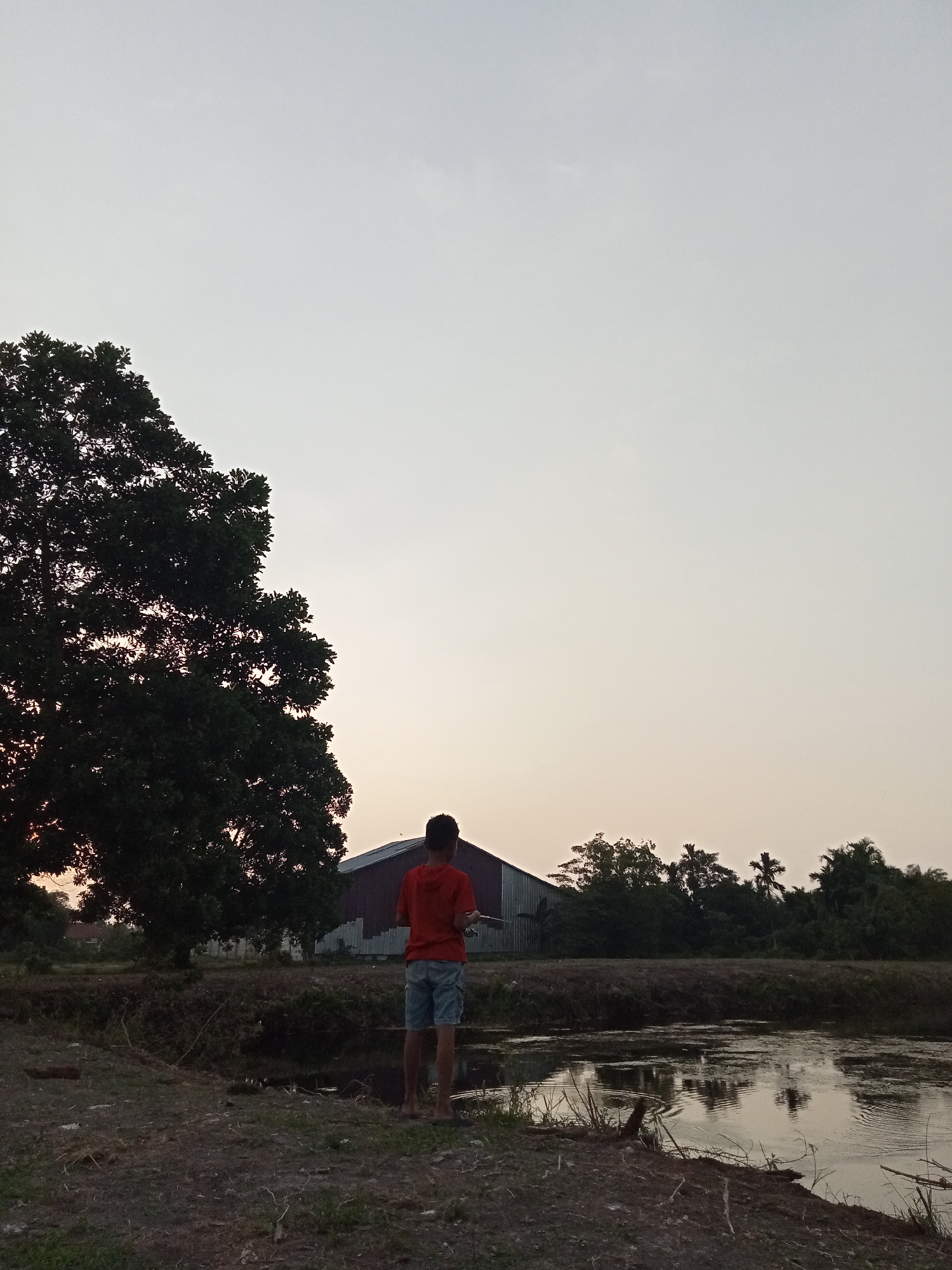 A young boy stands near the edge of a body of water, facing away from the camera. He is wearing a red hooded shirt and light-colored shorts, and appears to be holding a fishing rod. The water is calm, with reflections of the surrounding environment visible on its surface. Behind the boy, a large building with a metal roof is visible, as well as a dense area of trees. The sky is overcast and appears to be nearing sunset. The scene depicts a rural or semi-rural setting, possibly in Teluk Kenidai, Indonesia, with a focus on the boy's solitary activity by the water.