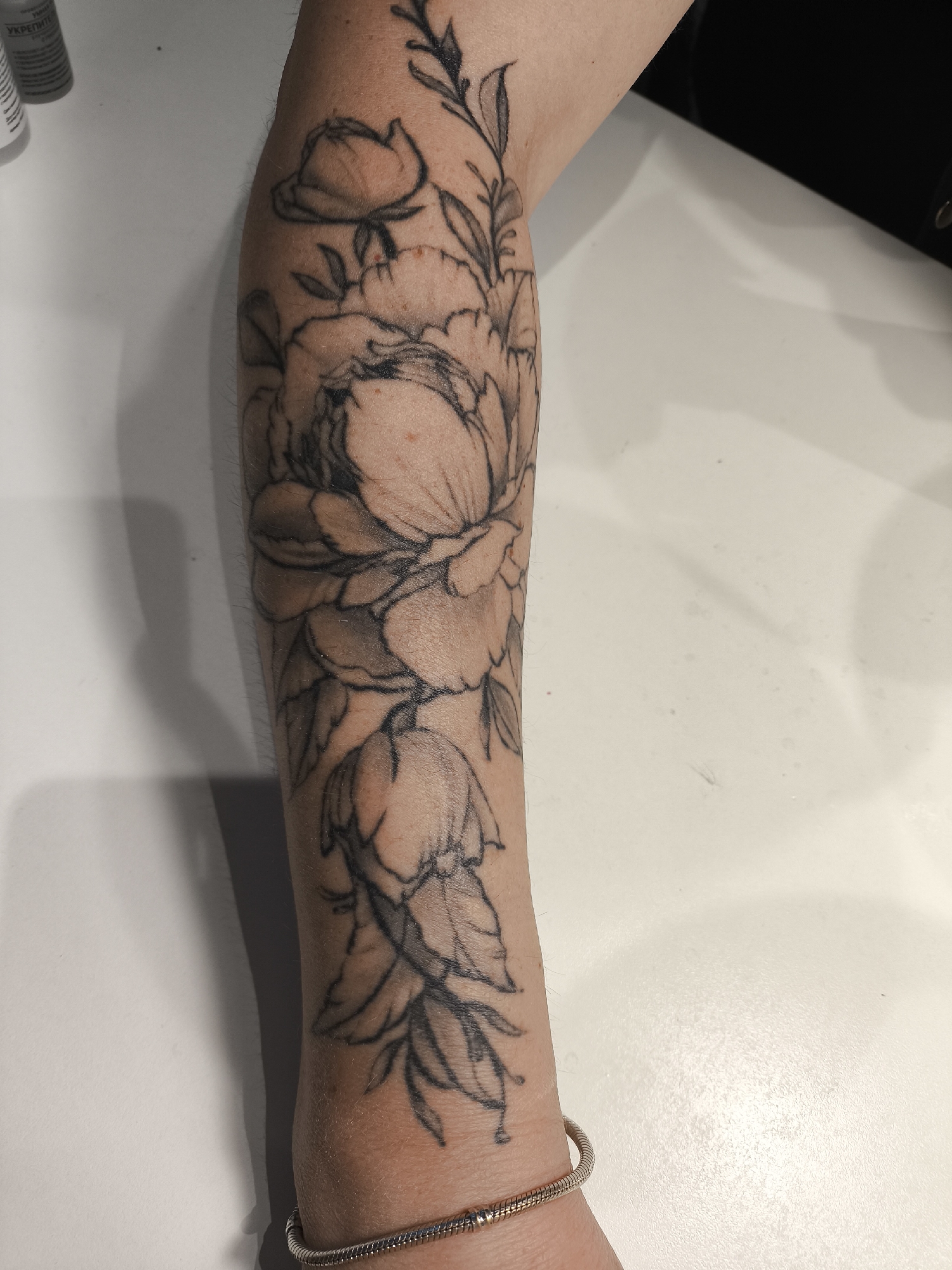 A person's forearm is shown from a top-down perspective, featuring a black and grey floral tattoo. The tattoo, which extends along the length of the forearm, depicts multiple flowers, including a prominent peony-like blossom in the center, surrounded by smaller buds and leafy branches. The design uses line work with light shading.
The skin of the forearm is light-toned, with some fine hair visible. A silver-colored bracelet with a segmented or beaded design is worn on the wrist at the bottom of the image.
The arm rests on a light-colored, flat surface, likely a table or countertop. A shadow is cast by the arm on the surface to its left. In the upper left corner, a portion of a white bottle is visible, bearing the partially legible Russian word "УКРЕПИТЕЛ" (UKREPITEL), which translates to "strengthener" or "fortifier." The scene is likely indoors, in Khimki, Russia.