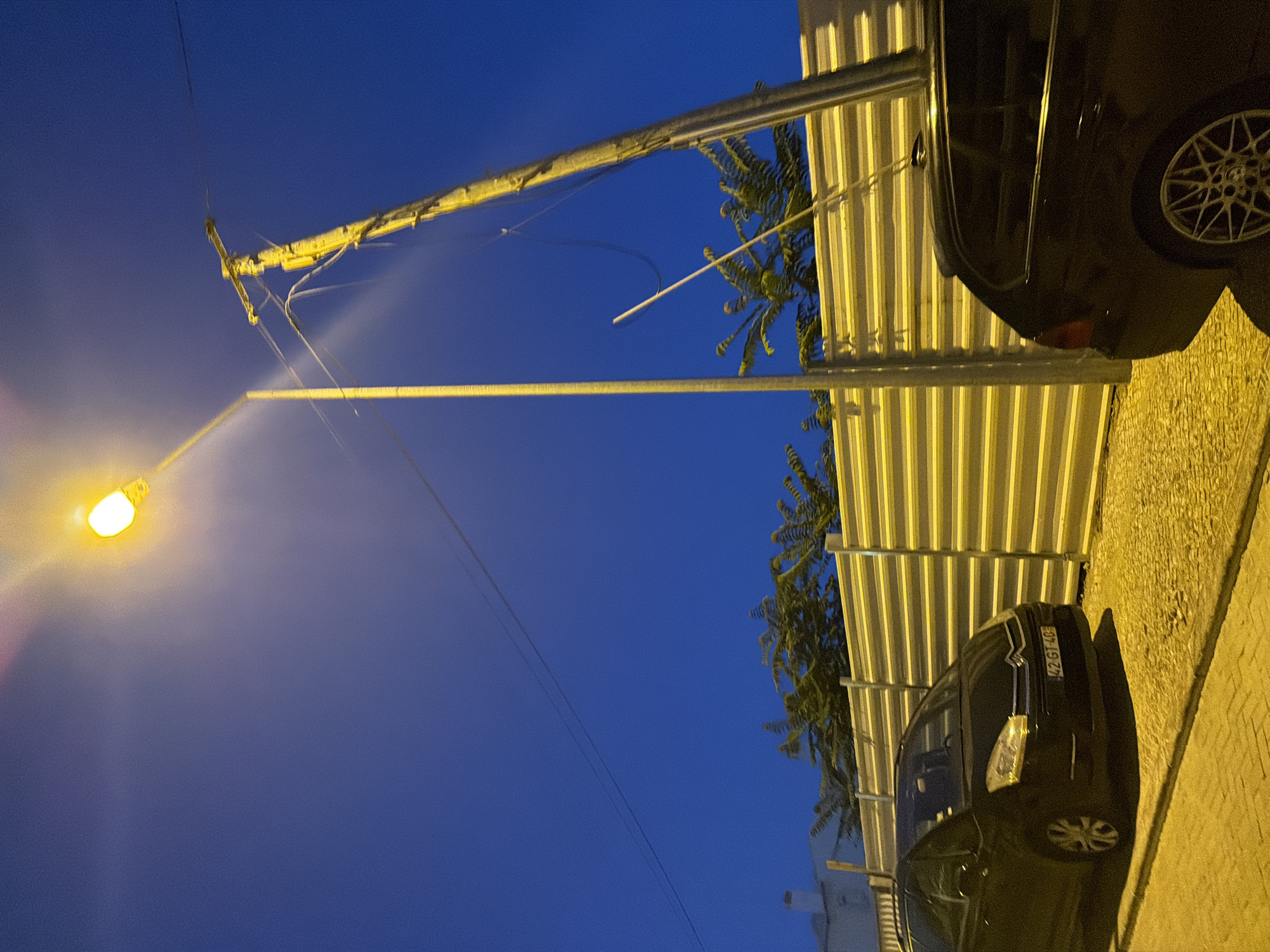 This image depicts a street scene at dusk or night in Cascais, Portugal. A bright street lamp illuminates the scene from the upper left quadrant, casting a warm glow. To the right of the street lamp, a wooden utility pole with wires extends upwards. In the foreground, two dark-colored cars are parked on a textured paved surface. The car on the left is a black Citroen minivan, identifiable by its license plate "42 GT 40", which is parked parallel to a yellow corrugated metal fence. Behind the fence, trees with feathery leaves are visible, and further back, a white building with a chimney can be seen. The sky is a deep blue, transitioning from the twilight hues near the lamp. The overall impression is one of a quiet urban street at the end of the day.
