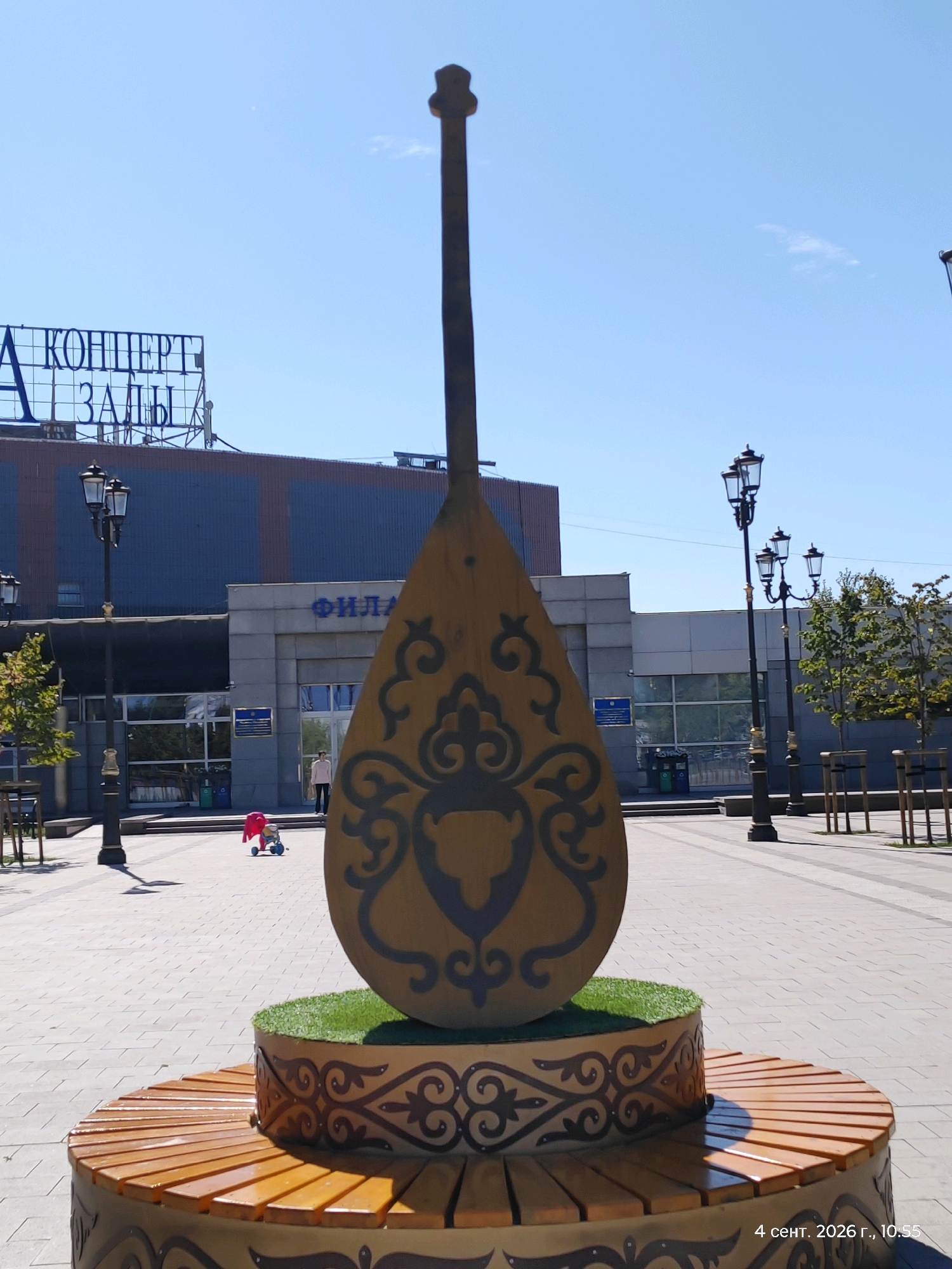 The image shows a large, stylized instrument, resembling a dombra, standing in an outdoor plaza in Astana, Kazakhstan. The instrument is made of wood and decorated with traditional patterns. It is mounted on a circular bench that also features decorative elements. In the background, a building with signs that read "КОНЦЕРТ ЗАЛЫ" (Concert Halls) and "ФИЛАРМОНИЯ" (Philharmonic) is visible. Several lampposts line the plaza, and a few people are present, including a child with a toy stroller. The ground is paved with interlocking bricks, and trees are planted along the side of the plaza. The sky is clear and blue. The timestamp at the bottom right corner indicates "4 сент. 2026 г., 10:55".