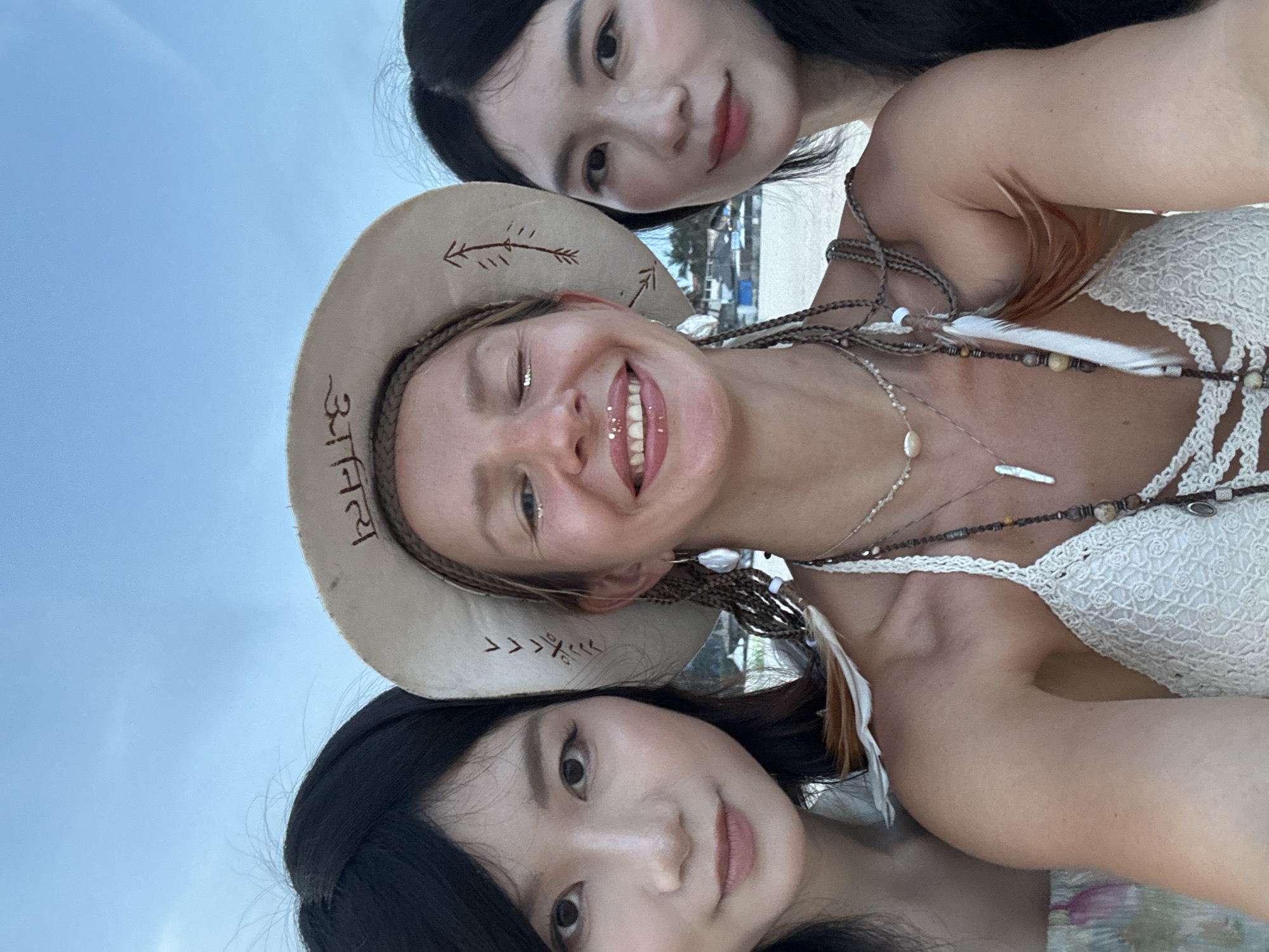 Three individuals, two women with dark hair and fair skin, and one woman with blonde hair, are positioned closely together, facing the camera. The blonde-haired woman, in the center, wears a wide-brimmed hat with an emblem and intricate beadwork and feathers adorning her hair and neck. She has glitter on her face and is smiling, with her mouth slightly open. The woman on the left has straight, dark hair and is looking directly at the camera with a subtle smile. The woman on the right also has dark hair and is looking towards the camera with a gentle expression. They are likely at a beach location, with a blurred background showing structures that could be buildings or huts, and the ocean or sky beyond. The image appears to have been taken outdoors, possibly during the day or late afternoon given the lighting. The context of Phu Thuy Ward, Vietnam, suggests a tropical or coastal setting.