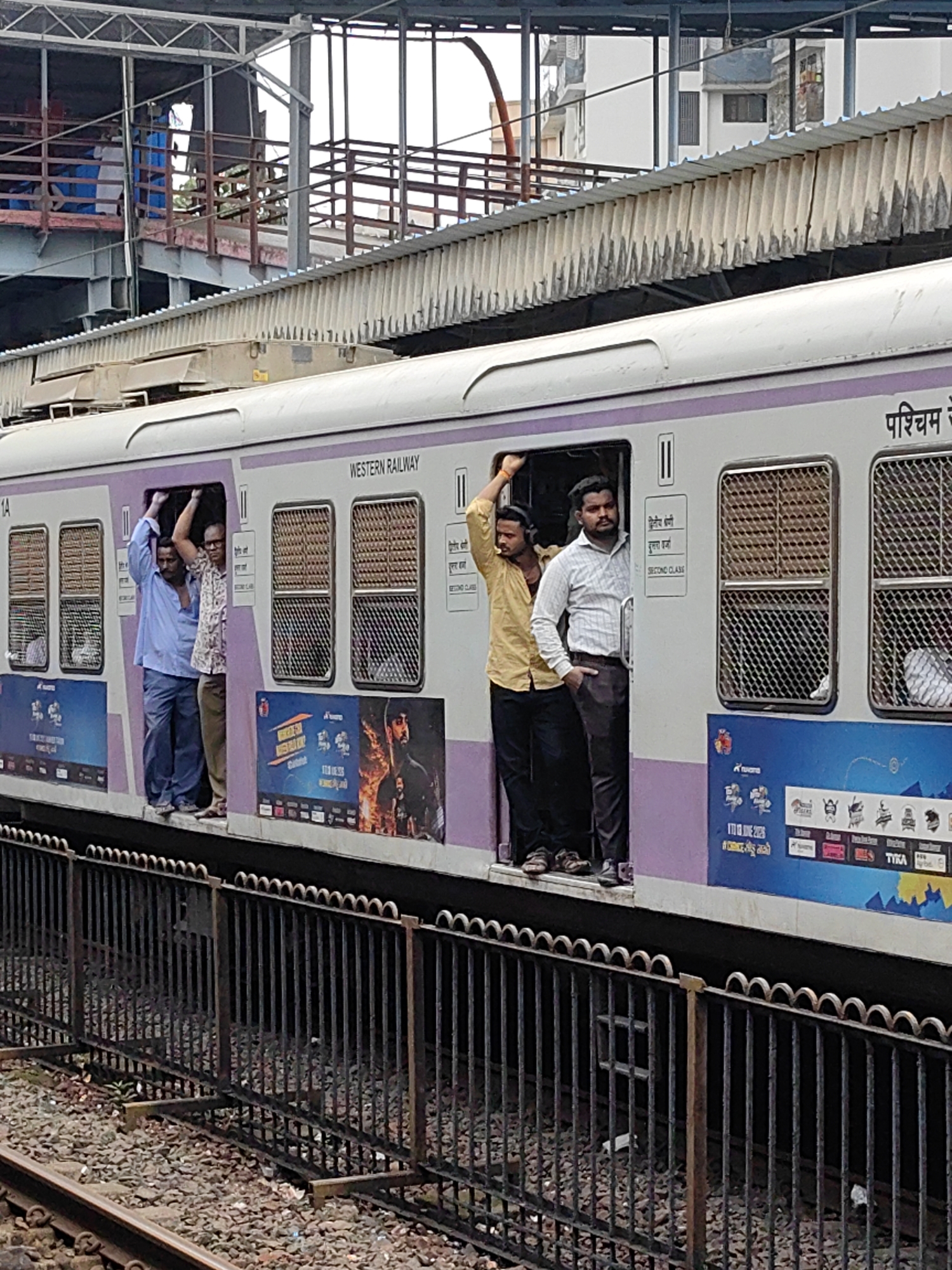 This is a medium shot, eye-level photograph of a crowded train car at a station in Mira Bhayandar, India. The train is light purple and white with "WESTERN RAILWAY" visible on its side. Several men are standing in the open doorways of the train, some with hands on the door frames, others with arms raised. One man is wearing a yellow shirt, another a light blue shirt and pants, a third a patterned shirt, and a fourth a light colored striped shirt and dark pants. The train is adjacent to a raised platform, visible in the background, which is covered by a corrugated metal roof supported by beams. A black metal fence runs along the edge of the platform in the foreground, separating it from the train tracks. Advertisements are visible on the side of the train car. The overall scene depicts a busy public transportation environment.