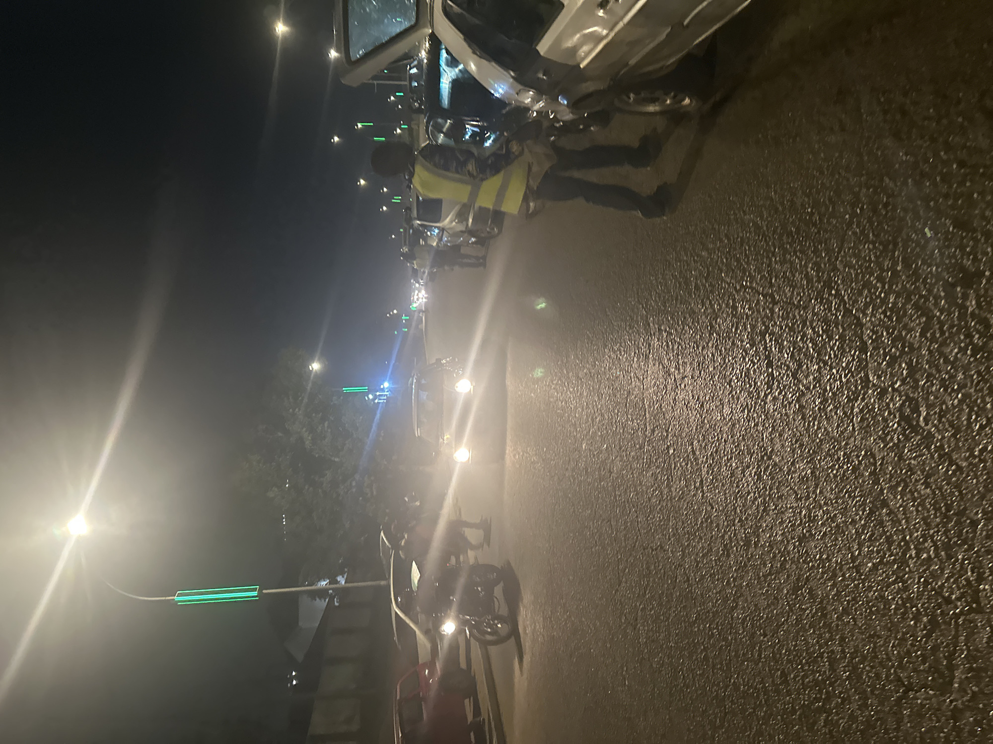 This is a night-time photograph taken on a wet asphalt road in Abuja, Nigeria. The scene depicts a traffic situation, possibly a checkpoint or a congested area. Several cars are visible, some parked and others moving. A motorcycle with its headlight on is positioned in the foreground on the left side of the image. A person wearing a high-visibility yellow vest and dark trousers is standing in the middle of the road, appearing to direct traffic or interact with a vehicle. Streetlights, some with green illuminated sections, line the road, casting light and creating reflections on the wet surface. The overall atmosphere is somewhat hazy, likely due to the lighting and the wet conditions. The background shows trees and the outlines of buildings, suggesting an urban or suburban setting.