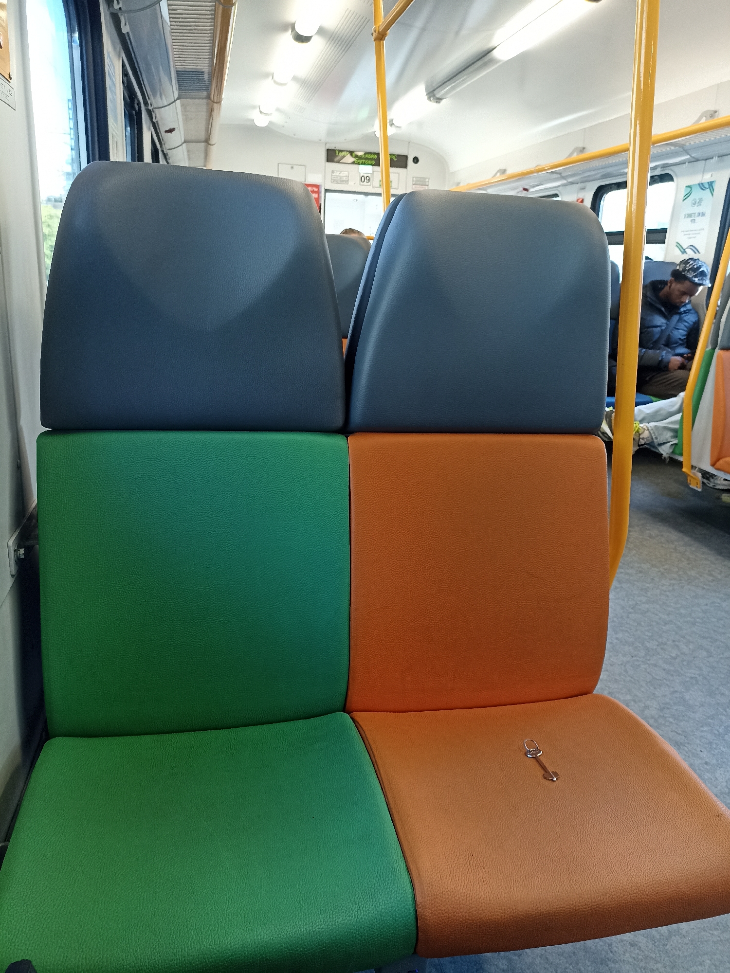 The image depicts the interior of a public transport vehicle, possibly a bus or train, with a focus on empty seats. The seats are a prominent feature, arranged in a 2x2 configuration in the foreground. The top two seats are dark blue, while the bottom two seats are a split design, with the left seat being green and the right seat being orange. A small silver key is visible on the orange seat in the foreground.
In the background, a person wearing a dark jacket and a patterned head covering is seated. They appear to be looking down at their phone. Yellow safety poles are visible throughout the carriage, a common feature in public transport. Advertisements or informational posters are also present on the walls. The lighting suggests it is daytime, with overhead lights on. The floor appears to be carpeted with a grey speckled pattern. The overall environment is clean and organized. There is no indication of the specific location within the city of Moscow, Russia.