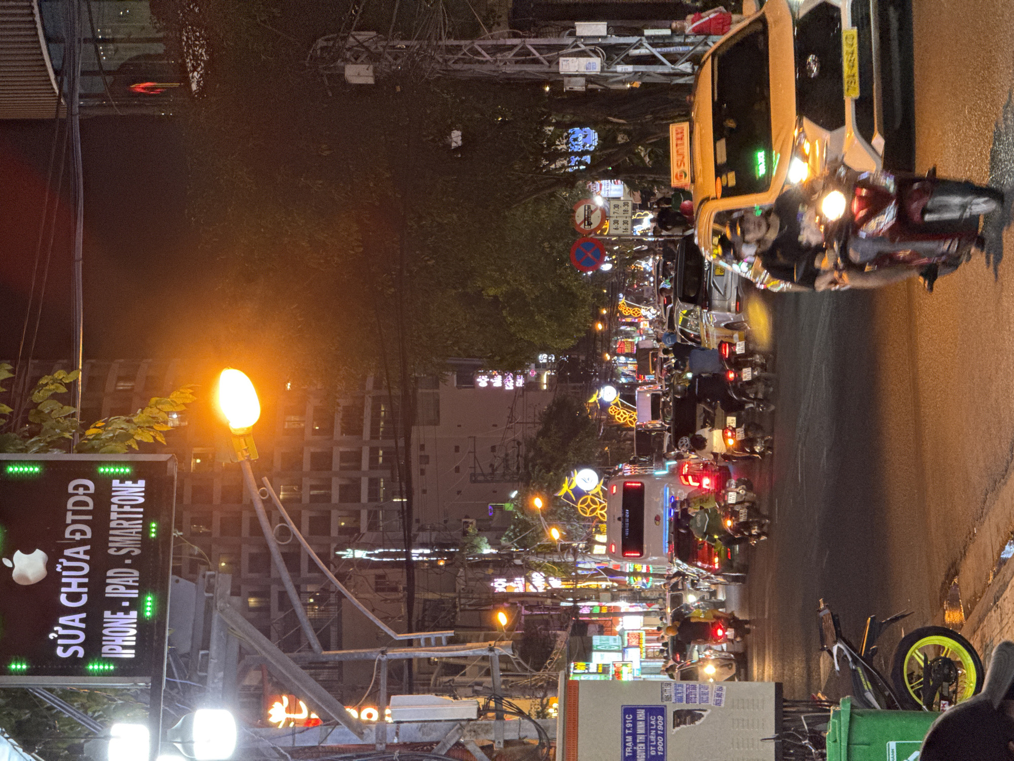 The image depicts a bustling city street scene at night in Nha Trang, Vietnam, likely along a main thoroughfare given the density of traffic and illuminated signage. The focal point is a bright LED sign displaying "SỬA CHỮA ĐTDĐ" (Phone Repair) in green letters, with an Apple logo above it and "IPHONE - IPAD - SMARTPHONE" below. Numerous motorcycles and cars are on the road, indicating significant traffic flow. A white bus is visible in the background, along with a yellow taxi with "SUN TAXI" branding. Several individuals are seen on motorcycles, some as drivers and others as passengers. Streetlights and neon signs of various colors illuminate the scene, reflecting off the wet asphalt, suggesting recent rain. The environment is characterized by urban infrastructure, including traffic signals, utility poles with visible wiring, and buildings lining the street, some with balconies and lighted windows. The overall impression is one of vibrant nighttime activity in a Vietnamese urban setting.