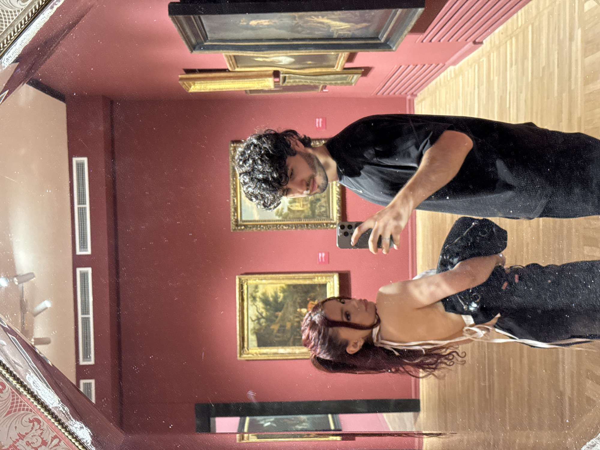 A young couple is taking a mirror selfie in what appears to be an art gallery. The woman, with long auburn hair tied in a ponytail, is wearing a white halter top and a dark skirt. The man, with curly dark hair and a beard, is wearing a black t-shirt. They are standing in front of several paintings displayed on a red wall. The floor is made of light-colored wood. The scene is illuminated by overhead lights. The image is captured from a low angle, showing the top of the ornate mirror frame in the foreground. The environment suggests it could be a museum or art exhibition. The couple's poses and expressions are casual, indicating they are enjoying their visit. There is no visible indication that this location is in Yerevan, Armenia, as the description is purely based on visual elements in the image.