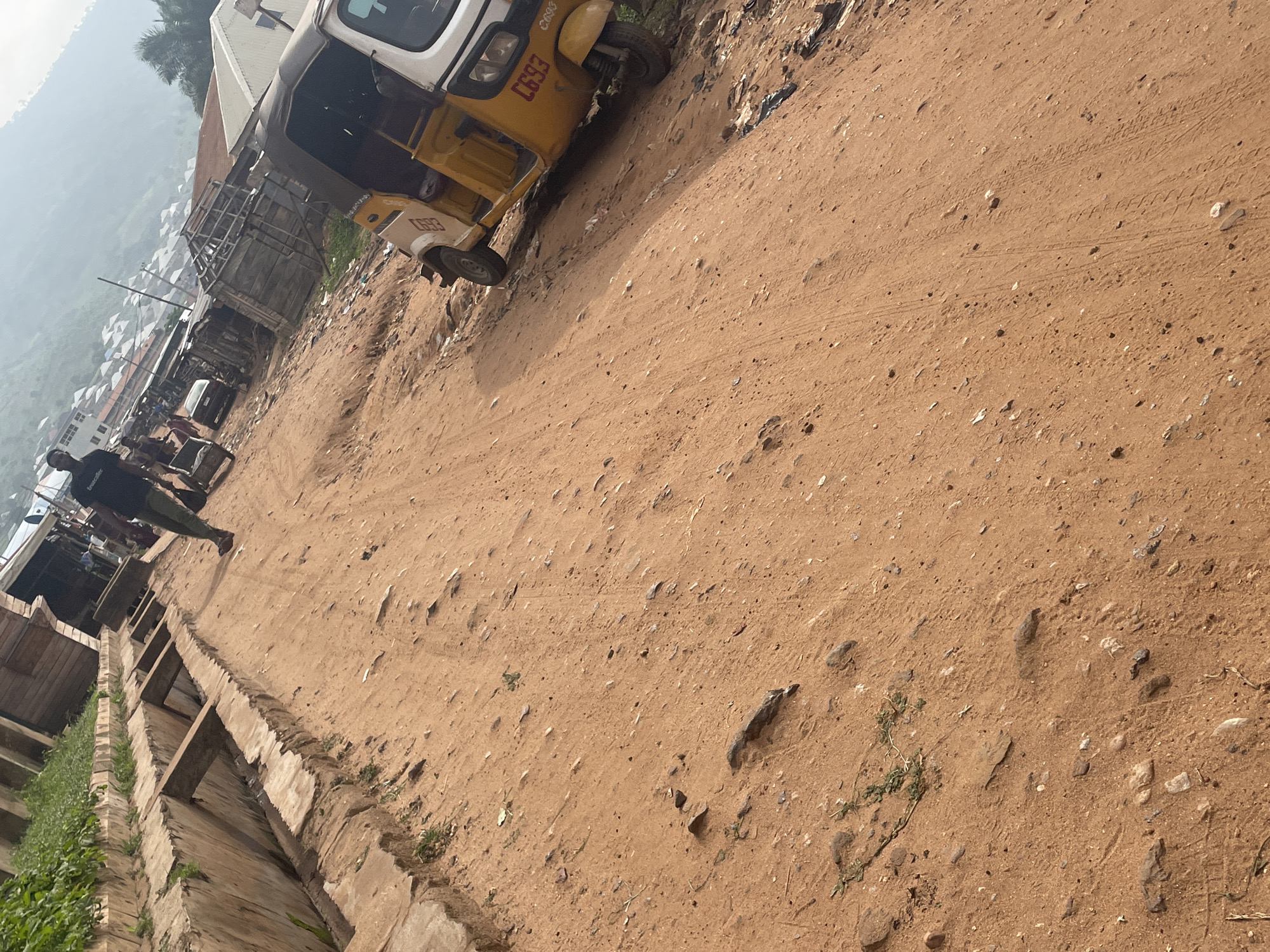 The image depicts a dusty, unpaved street in Lokoja, Nigeria. In the foreground, a yellow and white motorized tricycle, commonly known as an "Okada" or "Keke Napep", is parked on the right side of the road. Its license plate reads "LG93". A person in a dark t-shirt and khaki trousers walks away from the camera on the left side of the street. Behind them, a drainage ditch lined with concrete structures runs parallel to the road. The background reveals a densely populated urban area with numerous small buildings and structures, characteristic of a busy market or residential district in Lokoja. The overall scene conveys a sense of everyday life in a Nigerian city, with elements of transportation, infrastructure, and human activity.