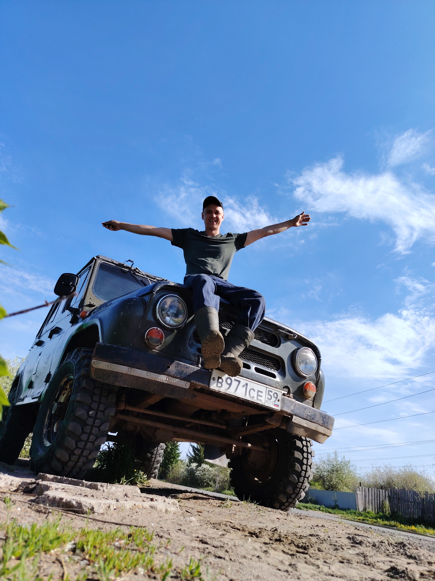 A low-angle shot captures a man seated on the hood of a dark green SUV against a bright blue sky with scattered clouds. The man, wearing a dark t-shirt, dark pants, and a baseball cap, has his arms outstretched, appearing cheerful. His boots rest on the front bumper of the vehicle, partially obscuring the license plate, which reads "B971CE 59 RUS." The SUV has large, rugged tires, indicating off-road capabilities. The vehicle is parked on a dirt and grassy surface.
In the background, a wooden fence and some low-lying bushes can be seen. Power lines are visible against the sky. The composition suggests a rural or semi-rural environment, not an urban scene. The image does not give enough information to definitively place the location in the city of Chaikovsky, Russia, and therefore the description is limited to what can be observed in the frame.