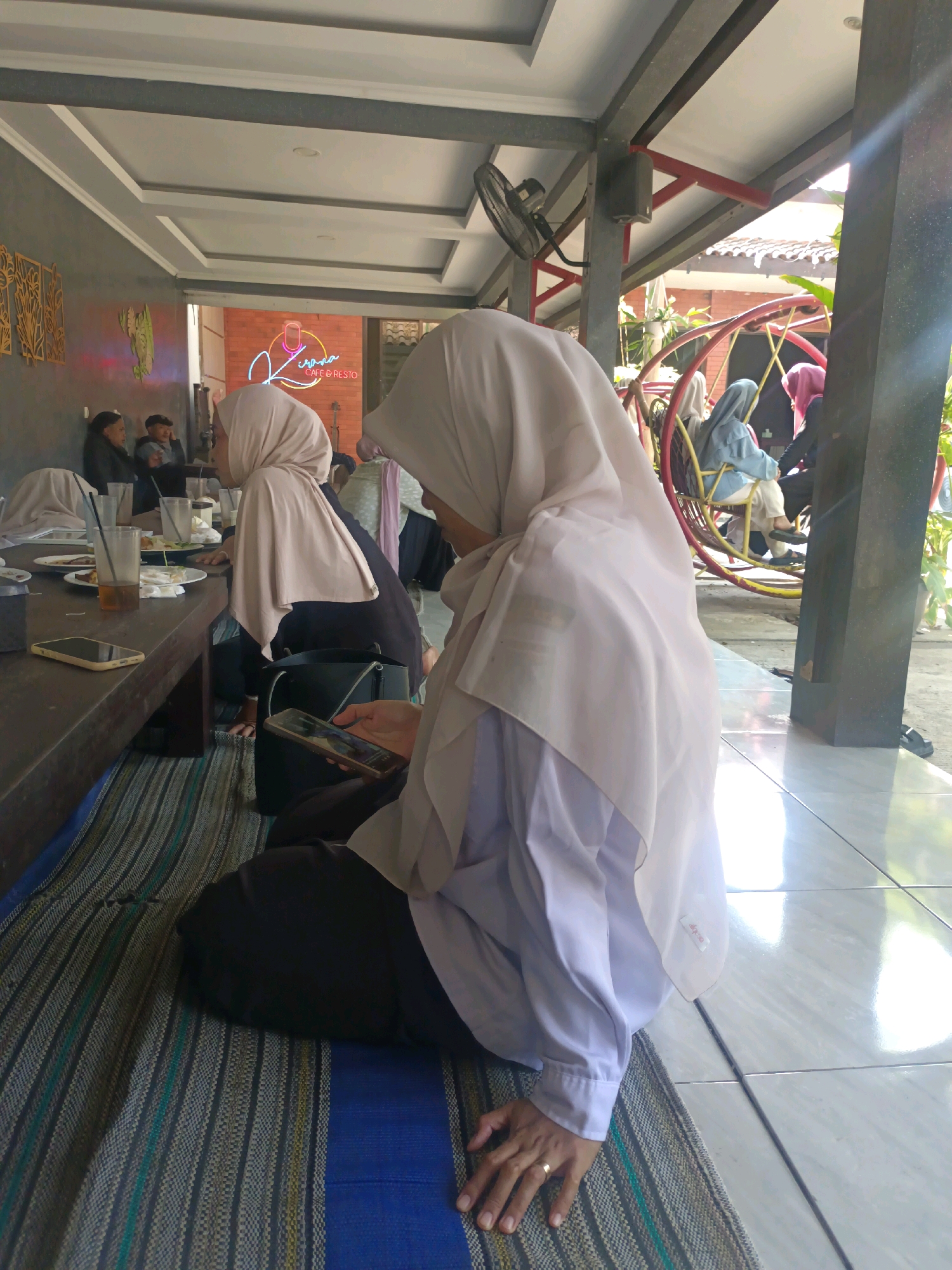 In Kerangkulon, Indonesia, a group of individuals are seated in a relaxed, open-air establishment with a shaded roof supported by dark pillars. In the foreground, a person wearing a light-colored hijab and a white shirt is seated on a patterned mat, looking down at a smartphone. Their left hand, adorned with a ring, rests on the mat. To their left, a wooden table holds various items including plates with food remnants, glasses with drinks, and a smartphone. Further back, other individuals are visible, some seated at the table and others on a yellow circular swing. The background shows a brick wall with a neon sign that reads "Jirapa CAFE & RESTO" and decorative wall art. The overall atmosphere appears casual and social.
