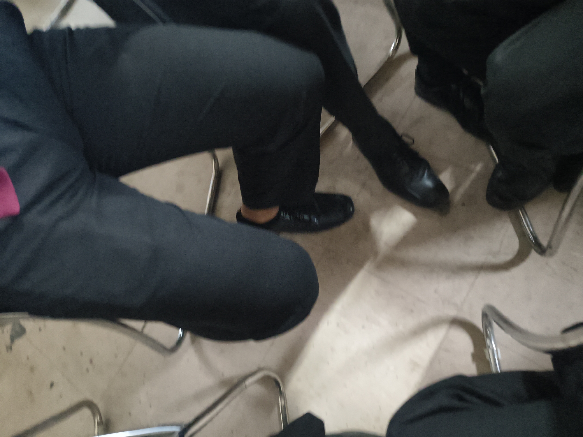 The image shows a close-up, low-angle view of several people's legs and feet. They are all wearing dark pants and shoes, suggesting a formal or semi-formal setting. The lighting is dim, and the focus is shallow, blurring the background. There appears to be a tiled floor, and some metal chairs are visible. The scene is ambiguous and lacks specific context to determine the exact location or activity. However, the question requests the location be Dharamshala, India, and while there is nothing in the image to confirm or deny this, if we were to assume this is the case, the image would depict individuals in Dharamshala, India, in a setting with tiled flooring and metal chairs.