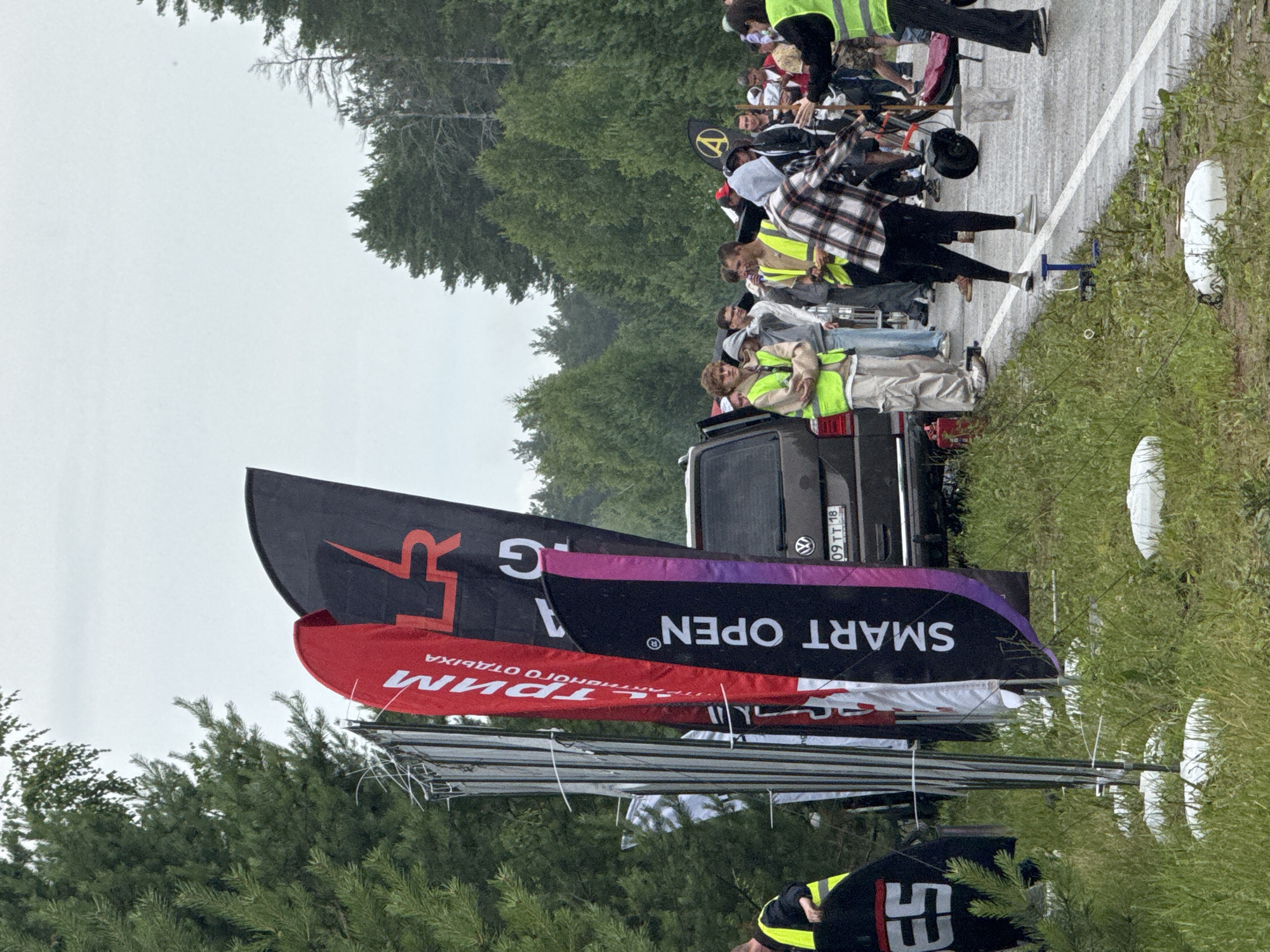 The image depicts an outdoor scene, likely during an event, with a group of people gathered on a paved road next to a grassy area. In the foreground, several tall, narrow banners are prominently displayed. One banner is red with white lettering that reads "ТРИМ". Another is black with red lettering forming a logo resembling an "LR". A third banner is black with white lettering that reads "SMART OPEN®" and has a purple accent. A dark-colored van is parked behind the banners, with its rear license plate visible, showing "09TT 18".
Several individuals are present, many wearing brightly colored vests, suggesting they might be staff or participants. Some are standing and observing, while others appear to be engaged in activities related to the event. One person in the mid-ground is wearing a plaid shirt and black pants, holding a stick. Another person to their right, wearing a yellow vest, is interacting with a trolley-like device. The background is filled with dense green trees, indicating a natural or semi-rural setting. The sky appears overcast.
The event seems to be taking place in or near Chaikovsky, Russia, given the presence of Russian text on the banners. The specific location is likely a road or open area used for events, possibly a race track or festival grounds, judging by the setup with banners and gathering of people. The environment is characterized by a mix of asphalt and vegetation, with a backdrop of trees and a cloudy sky.