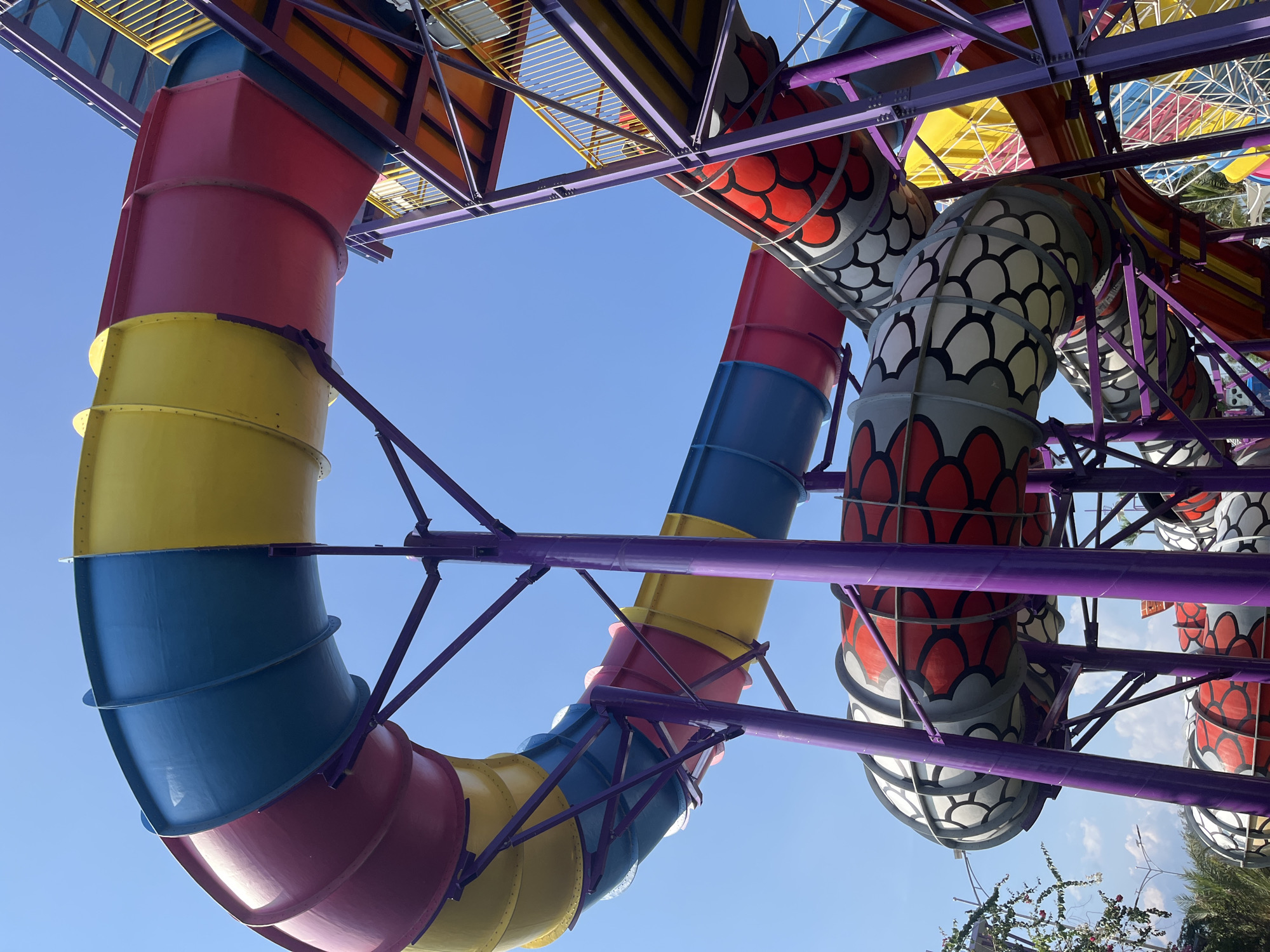 This image captures a vibrant water slide structure against a clear blue sky in Dong Ninh Hoa Ward, Vietnam. The slide is designed with large, winding tubes in a variety of colors, including red, yellow, and blue, with segments resembling a dragon's scales. The structure is supported by a sturdy purple metal frame. Multiple levels and platforms are visible, indicating a complex and multi-faceted water attraction. The lighting suggests it is daytime, with bright sunlight illuminating the scene. No people are clearly visible in the image.