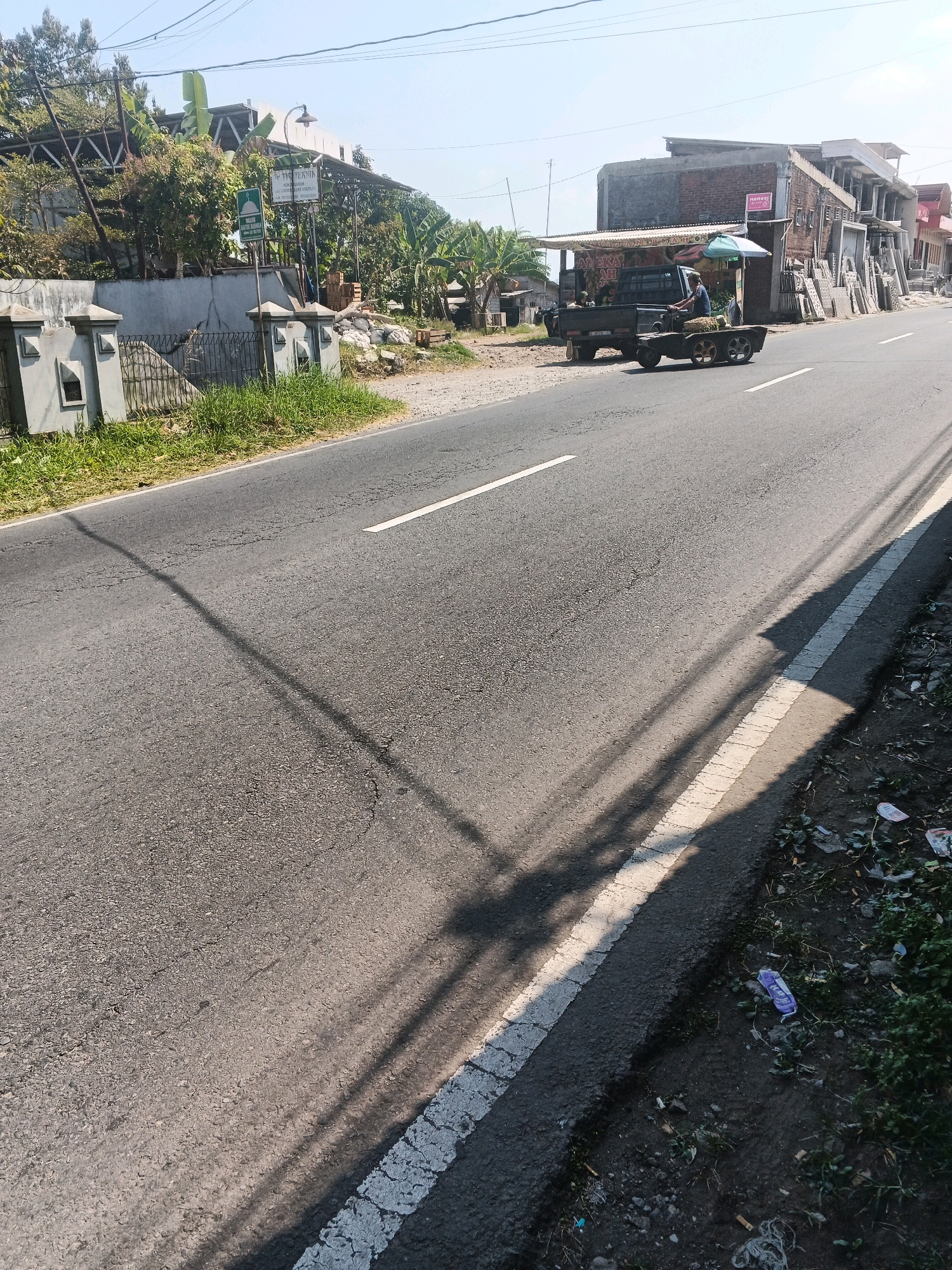 In Bandungan, Indonesia, a road scene unfolds under a clear sky. On the left side of the asphalt road, a low concrete wall with decorative pillars and a metal fence marks the boundary of a property. Beyond the wall, lush green grass grows, and trees and buildings with visible roofing structures are present. A green sign with a mosque symbol and Indonesian text is mounted on a post. Further along the road, a dark-colored pickup truck is parked. A person is seated in the driver's seat of the truck, which is towing a trailer filled with what appears to be agricultural produce covered by a green umbrella. In front of the truck, a small building with a corrugated roof and advertisements is visible, and a collection of stacked materials, possibly building supplies, lines the roadside. On the right side of the road, the asphalt surface shows signs of wear and cracking, with a white line marking the edge. Debris, including plastic wrappers and cigarette butts, litters the unpaved shoulder of the road, which is bordered by vegetation. The scene is bathed in sunlight, casting long shadows from utility poles and trees across the road.