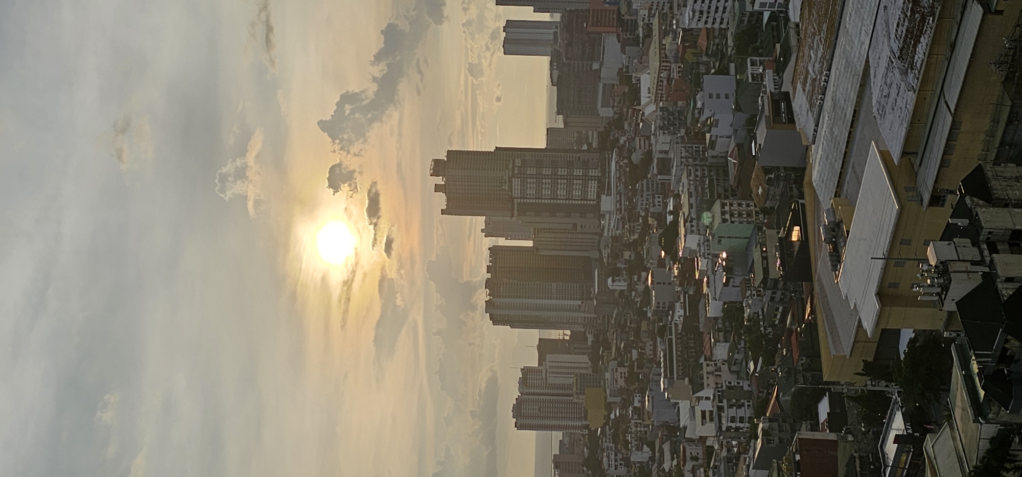 This is a vertical, high-angle, aerial shot of a city skyline in Makati, Philippines, during sunset or sunrise. The sun, a bright white orb, is positioned in the upper center of the frame, casting a warm, golden light across the sky and the cityscape. Clouds, ranging from wispy and translucent to more solid and gray, are scattered throughout the sky, some illuminated by the sun.
The cityscape is densely populated with a mix of modern high-rise buildings and lower-rise residential or commercial structures. Prominent among the tall buildings are several skyscrapers with a contemporary architectural style, characterized by numerous windows and geometric shapes. In the middle ground, a sprawling network of smaller buildings with varied rooflines and colors fills the space between the towering structures. The lower foreground reveals more of these smaller buildings, some with visible rooftops, air conditioning units, and satellite dishes. A hint of a body of water, possibly Manila Bay, can be seen in the far distance beyond the buildings. The overall atmosphere is one of a bustling urban environment bathed in the soft glow of the setting or rising sun. There are no visible people in the frame.