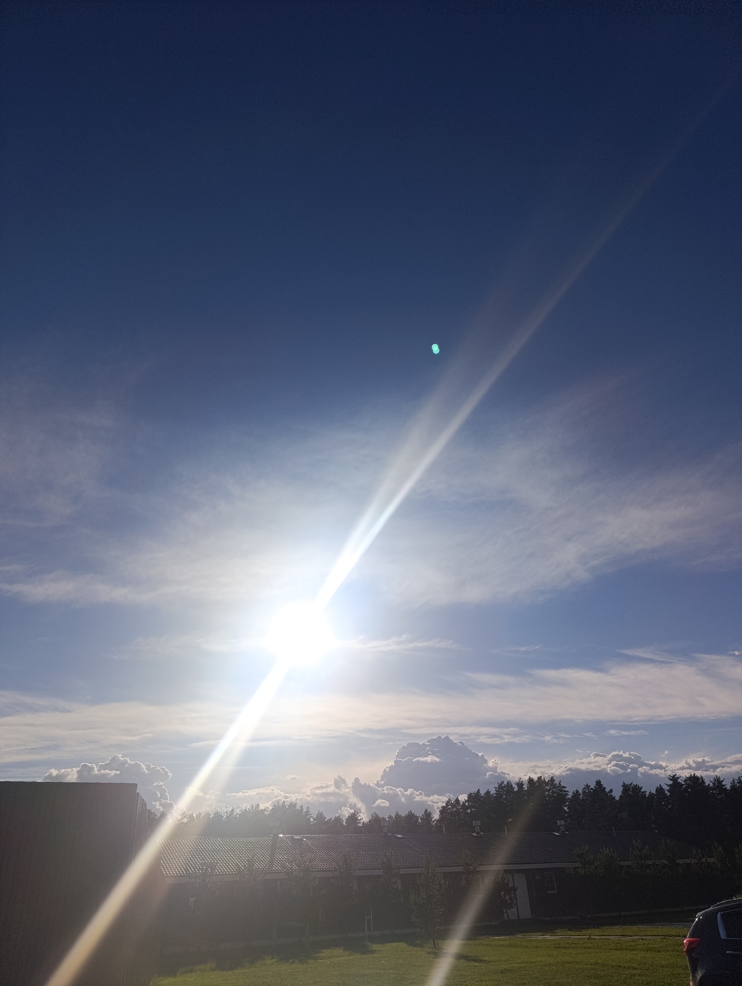 The image shows a bright sun in a blue sky with some scattered clouds. A prominent light flare extends downwards from the sun. In the foreground, there is a building with a dark roof and a grassy lawn. A line of trees is visible behind the building. A portion of a car is visible in the bottom right corner of the image. The location is Unknown, Unknown.