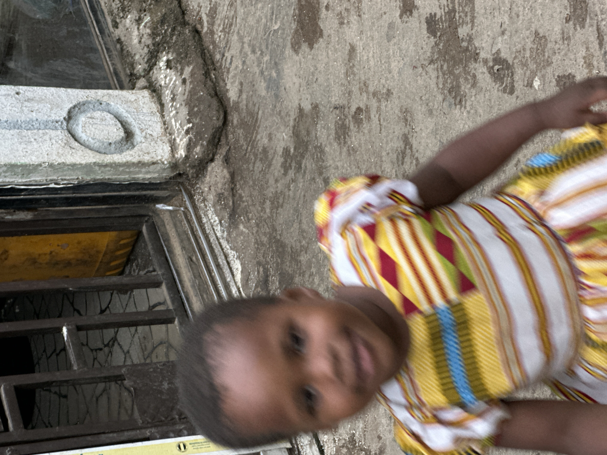 The image is a medium shot of a young Black child, appearing to be a girl, wearing a brightly colored, patterned dress. The dress has a white base with horizontal stripes of yellow, red, green, and blue. The child is in motion, which causes a blur in the image, particularly around her face and body. She has dark hair and her facial features are somewhat indistinct due to the motion blur. The child is outdoors, with a textured, grayish concrete ground covering the lower portion of the frame. In the background, there is a dark metal gate with vertical bars, and beyond the gate, a tiled floor can be seen. To the right of the gate, a textured concrete wall is visible, with a circular indentation or marking on its surface. Behind the wall, a blurry window is present, and a yellow object, possibly a container or barrel, is partially visible through the gate. The overall environment suggests an urban setting.