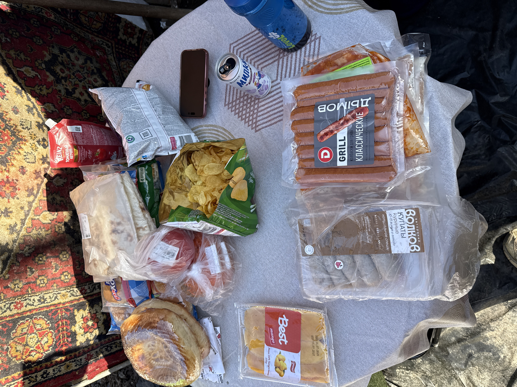 On a light-colored tablecloth spread over a surface, possibly a table, rests an array of food items. A blue sports bottle and an open can of “LOTTE Милк” stand near a smartphone. Visible food items include two packages of sausages, one labeled "Дымов GRILL Классические" and the other "Волков Купаты". A bag of potato chips, a package of "Best mango" dried mango slices, tomatoes, and bread are also present. A pouch of Heinz tomato ketchup and a package of buns are visible near a carpet with a red and black pattern. The environment suggests an outdoor setting, potentially a picnic or a meal being prepared outdoors. It's impossible to identify the exact location as Krasnoyarsk, Russia, only based on the image.