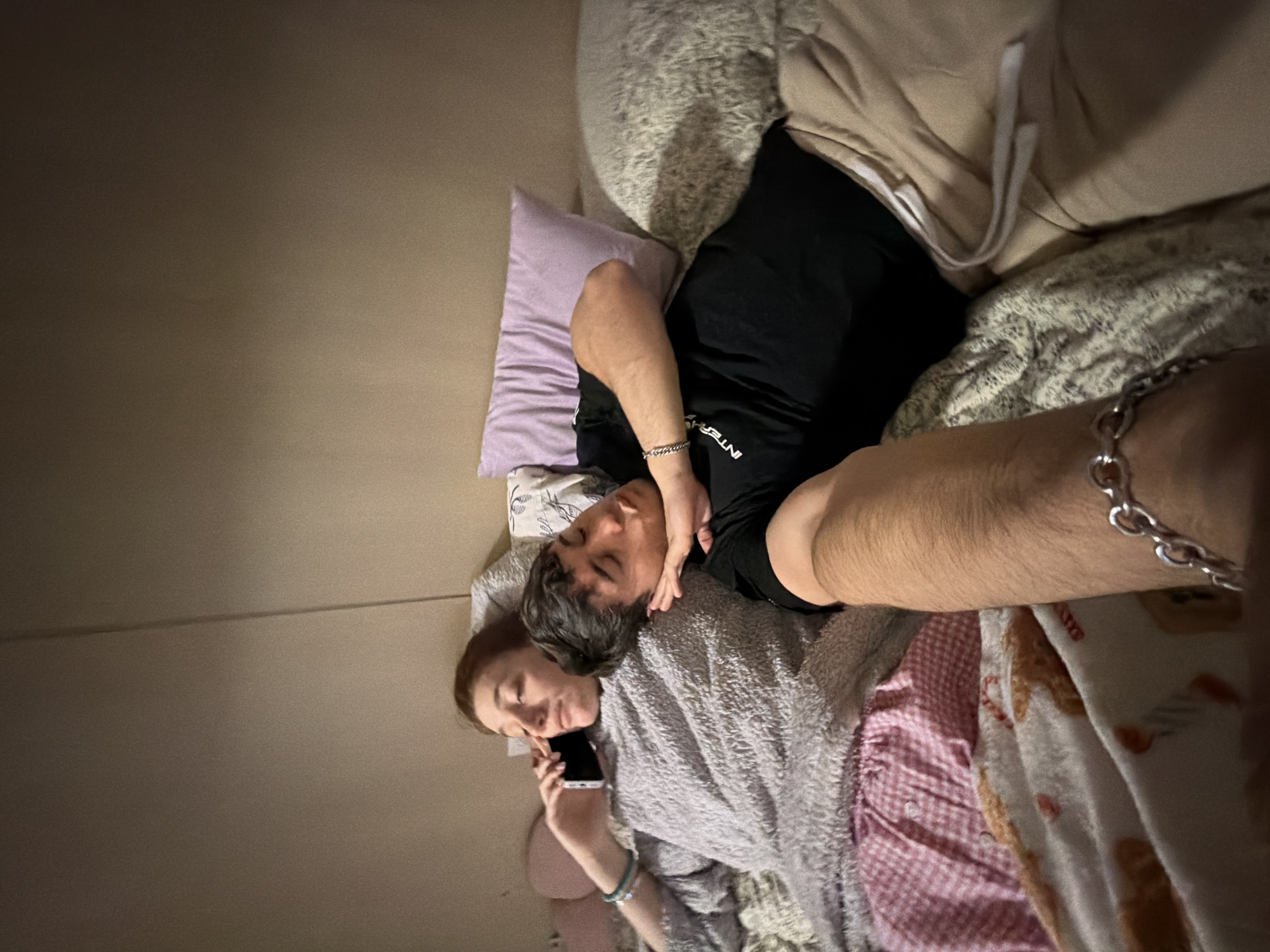 In an indoor setting, likely a bedroom in Sofia, Bulgaria, two individuals are seen lying on a bed in dim lighting, suggesting evening or night. The overall atmosphere appears peaceful and intimate.
On the right side of the frame, a male with dark hair lies on his side, his head resting near the female's shoulder or chest. He is wearing a black t-shirt, with the partial word "INTERP" visible in white text on it. A silver chain bracelet adorns his right wrist. His eyes appear closed, indicating he might be resting or sleeping.
To the left, a female with reddish-brown hair is lying on her back, holding a dark smartphone to her right ear, seemingly engaged in a phone call. She is covered by or wearing a soft, grey textured material, possibly a blanket or a plush robe. A light blue/green beaded bracelet is visible on her left wrist. Her expression is calm and focused on the call.
The bed is furnished with various bedding items, including a light purple pillow, a fluffy light grey pillow, a pink checkered blanket, and a white patterned blanket on the far right. The wall in the background is a plain light brown/beige color.
