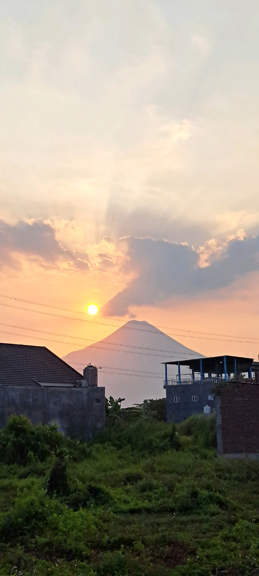 The image captures a serene sunset scene in Kemiri Sewu, Indonesia, with a prominent mountain silhouetted against the warm, golden hues of the sky. The sun, a bright orb, is partially obscured by scattered clouds, casting rays of light that pierce through the atmospheric haze. In the foreground, a residential area is visible, featuring a tiled-roof house on the left, a multi-story building with a blue-railed balcony in the center, and a brick structure on the right. Lush green vegetation covers the ground, creating a natural contrast with the man-made structures. Power lines crisscross the scene, adding a touch of utility to the otherwise natural landscape. The overall impression is one of tranquility and the beauty of nature coexisting with human habitation.
