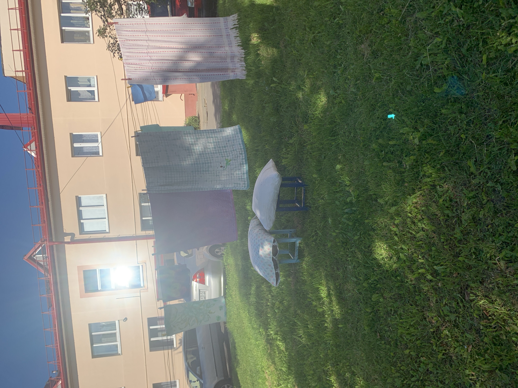 In Kapyĺ, Belarus, on a sunny day, laundry hangs on a clothesline in front of a light beige apartment building with a red roof. Several pieces of fabric, including a patterned pink and white cloth and a light blue cloth, are visible. In the foreground, two small stools with cushions sit on a patch of green grass. A silver car is parked behind the hanging laundry. The overall scene suggests a domestic setting with laundry being air-dried outdoors.