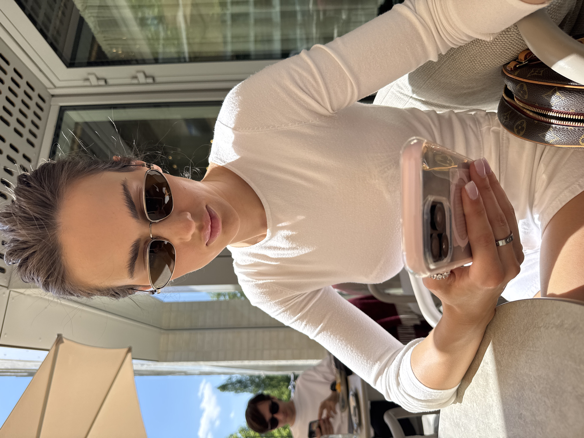 A woman wearing sunglasses is sitting at a table, holding a smartphone. She is wearing a white long-sleeved shirt and a ring on her left hand. Part of a brown handbag with a logo is visible to her right. In the background, another person is seated at a table, also wearing sunglasses. The setting appears to be an outdoor patio or café, with a building visible in the background, featuring a window with a shutter-like design and an awning.