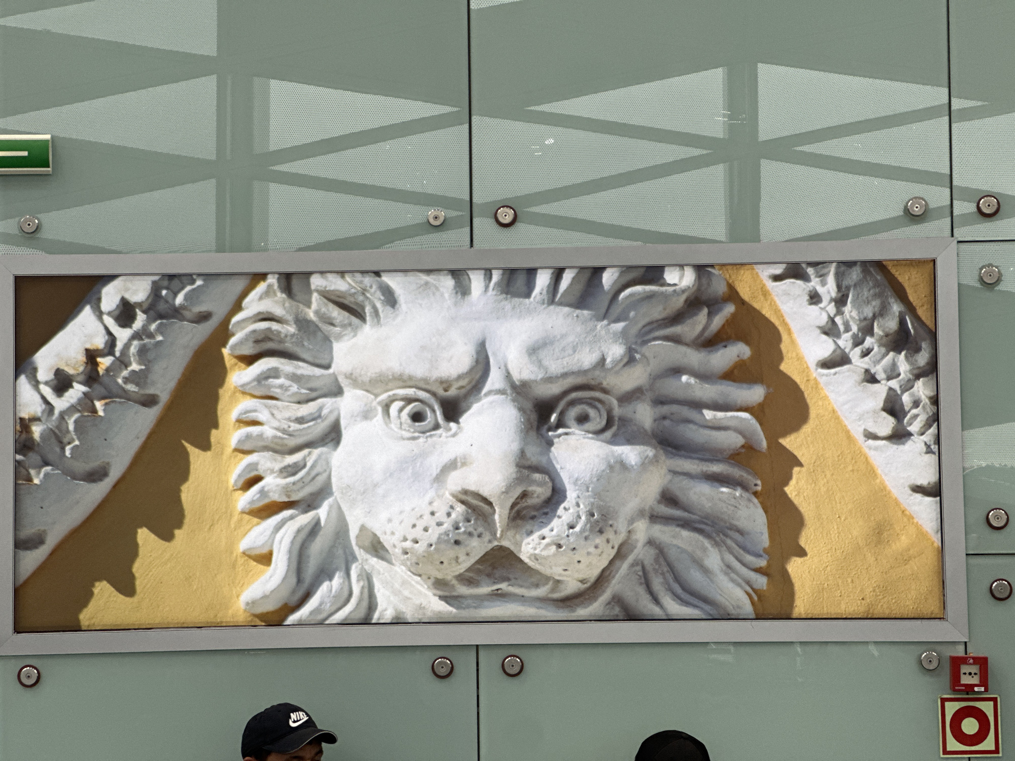 A close-up shot captures a bas-relief carving of a lion's face set within a rectangular frame. The carving is primarily white, with a textured mane and expressive eyes, set against a yellow background. The frame appears to be metal and of a pale color. The carving is mounted on a wall composed of large, opaque glass panels secured with visible metal bolts. The glass panels display a geometric pattern of translucent lines. At the upper left of the scene, a green emergency exit sign is visible. Below the carving are fragments of two individuals wearing dark clothing and caps, only their heads are visible. A red fire-alarm device is mounted on the wall below the bas-relief's frame. The architecture and aesthetic suggest the scene is likely located in Saint Petersburg, Russia.