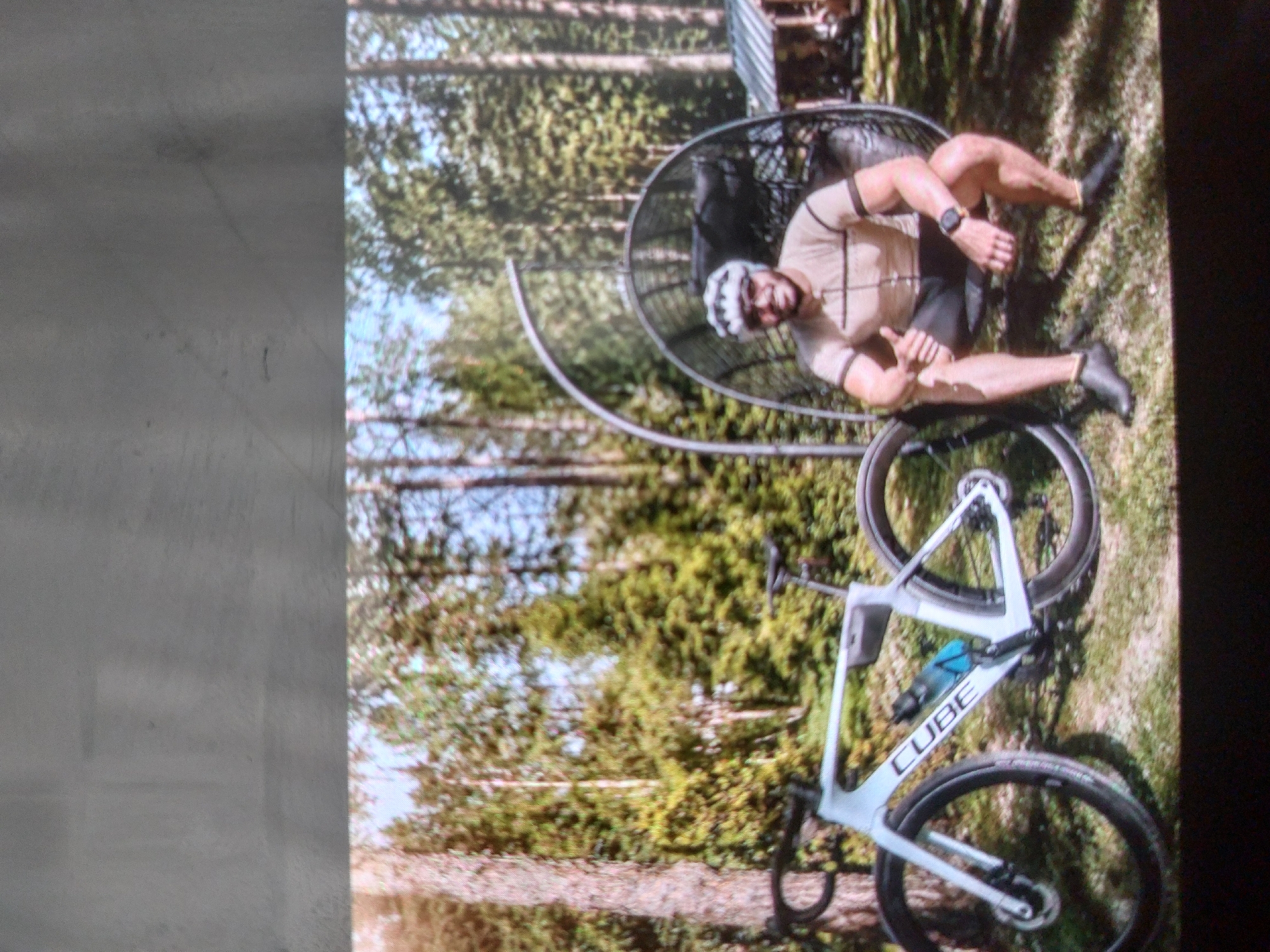 A cyclist wearing a helmet and sportswear sits in a chair with a bicycle next to him. The cyclist is in a forest setting. No specific location details for Tsarevo, Bulgaria are discernible.