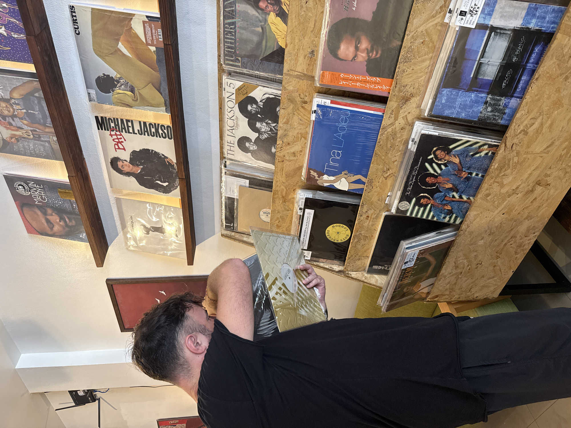 A person wearing a black t-shirt and dark blue shorts is looking through a wooden display rack filled with vinyl records in a shop. The rack is positioned in the foreground, and the person is bending over, holding a gold-colored vinyl record in plastic packaging. Behind the person, a white wall with some electrical equipment is visible. Above, a wooden shelf displays several vinyl records, including ones featuring Marvin Gaye, Michael Jackson's "Bad," and The Jackson 5. Further back, another shelf holds a record with the name "CURTIS" and another with "LUTHER VAN." The shop appears to be well-lit, with warm lighting from above. The overall scene suggests a record store environment. The image is undated and lacks specific location indicators within the city of Ko Samui District, Thailand, beyond the inferred context of a shop.