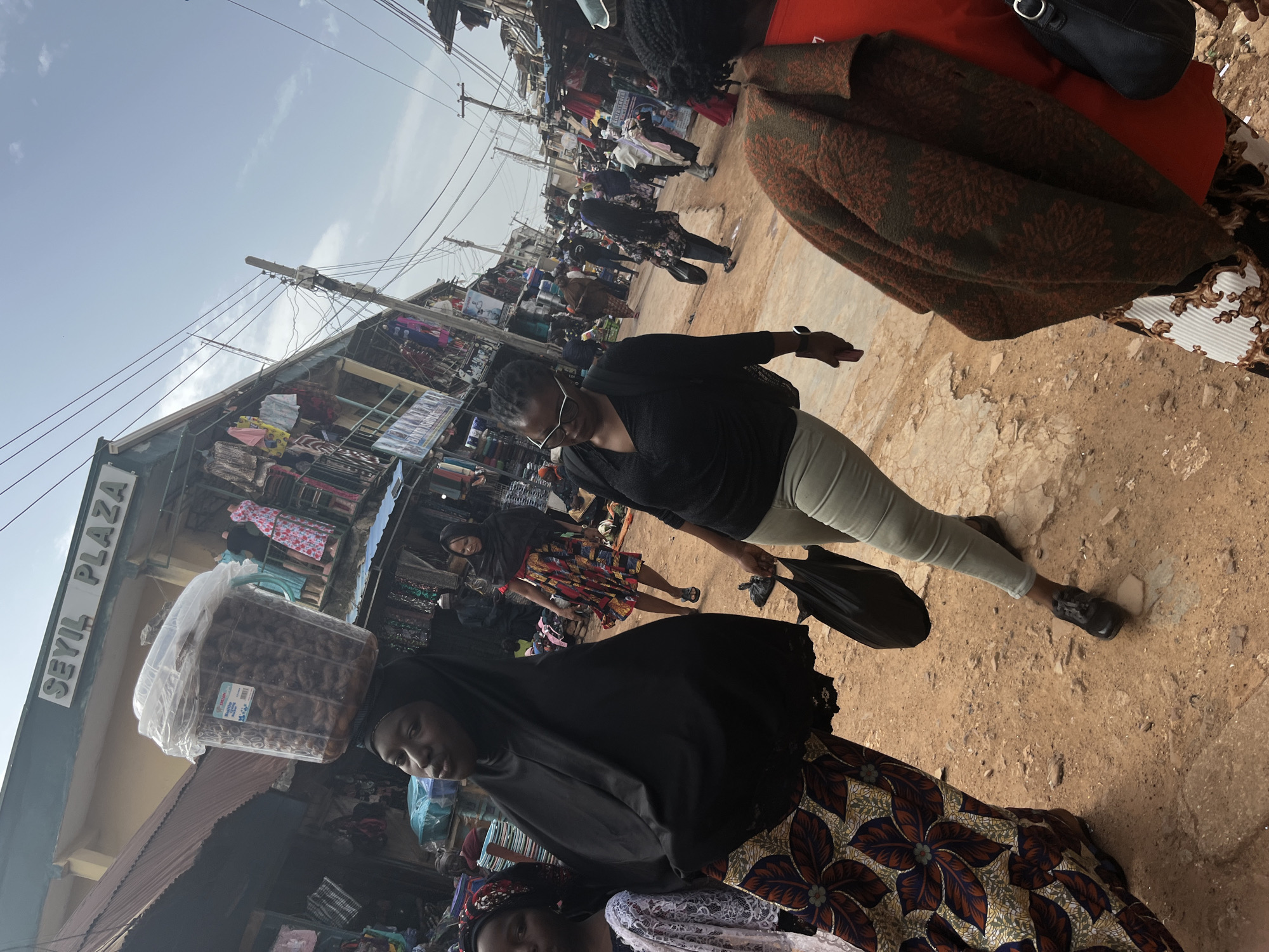 The image captures a bustling market scene in Jos, Nigeria, specifically outside Seyil Plaza. In the foreground, a woman wearing a black hijab and a vibrantly patterned wrap skirt carries a large, clear plastic container filled with dark, dried items on her head. To her right, another woman, in the mid-ground, walks away from the camera wearing glasses, a black top, and light-colored trousers, carrying a black plastic bag. Behind them, the market extends with stalls displaying clothing and goods. The ground is unpaved, consisting of dirt and scattered debris. The overall impression is of a busy commercial environment under a bright, partly cloudy sky, with numerous people navigating the crowded pathways between shops.