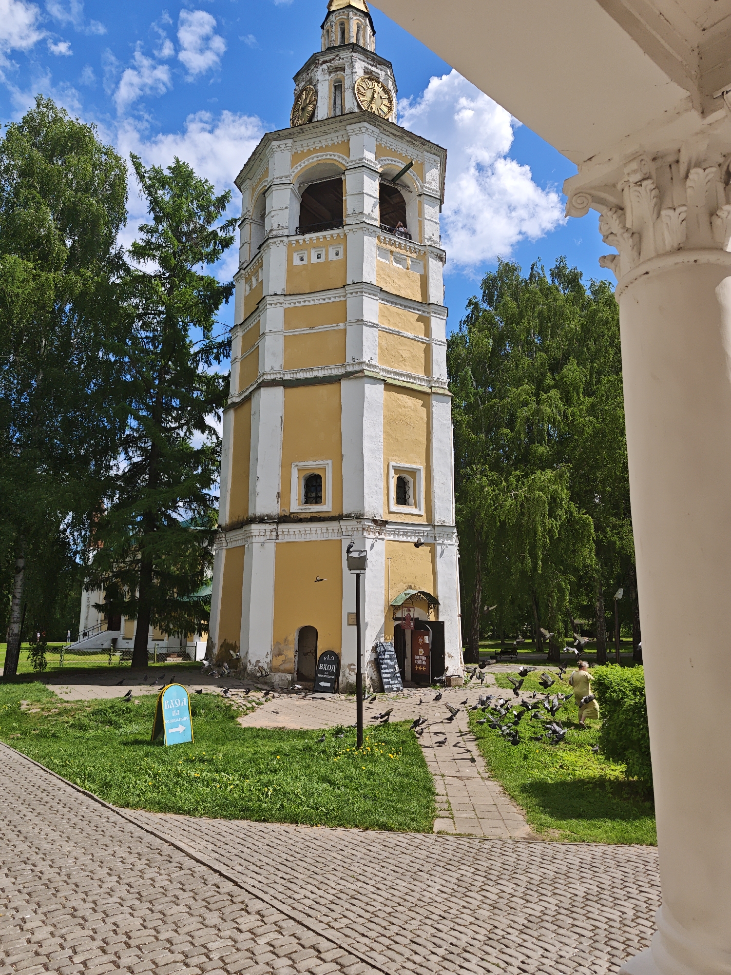 The image shows a yellow and white bell tower in what appears to be a park. The bell tower has a clock at the top and several tiers. In front of the tower, there is a group of people feeding pigeons. The scene is shot on a sunny day with a blue sky and white clouds. The image is taken from a low angle, and a white column is partially visible in the foreground. A sign is also visible, but the text is not clearly readable. Based on the architecture, the location is in Suzdal, Russia.