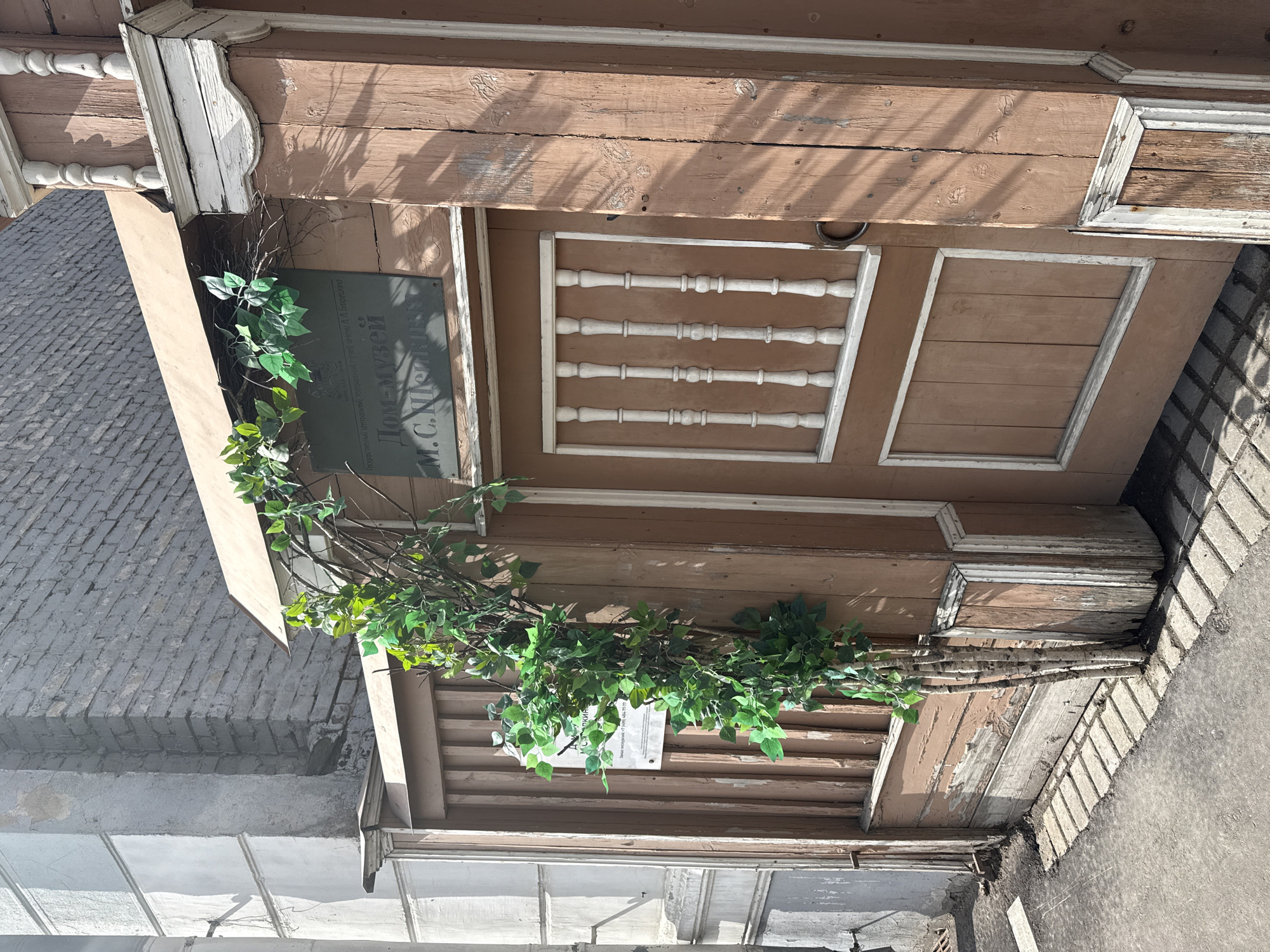 The image shows the exterior of the M.S. Schepkin House-Museum in Moscow, Russia. The building is constructed of brown wood, with decorative white trim. A green vine adorns the facade, partially obscuring a sign. The building is adjacent to a brick wall and concrete sidewalk. The sky is visible in the background.
