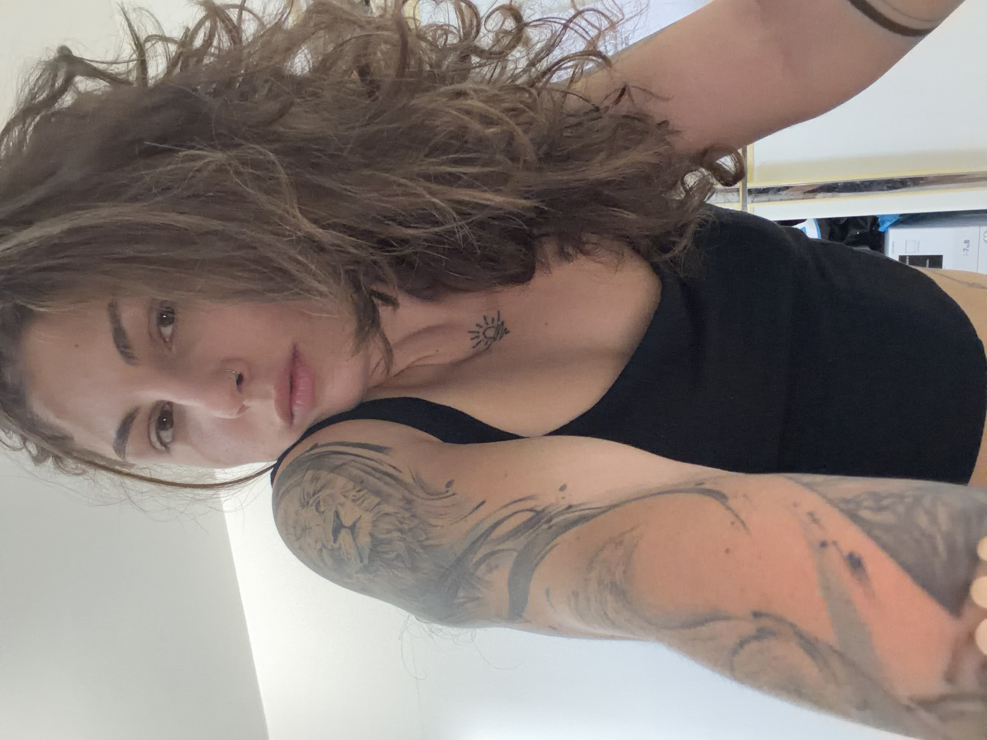 A woman is in the image. She has long, curly brown hair, a fair complexion, and tattoos on her left arm and chest. She is wearing a black tank top. The background is a white wall, with a glimpse of a white shelf or cabinet visible. The image provides no information to determine its location in the city of Kiryat Bialik, Israel.