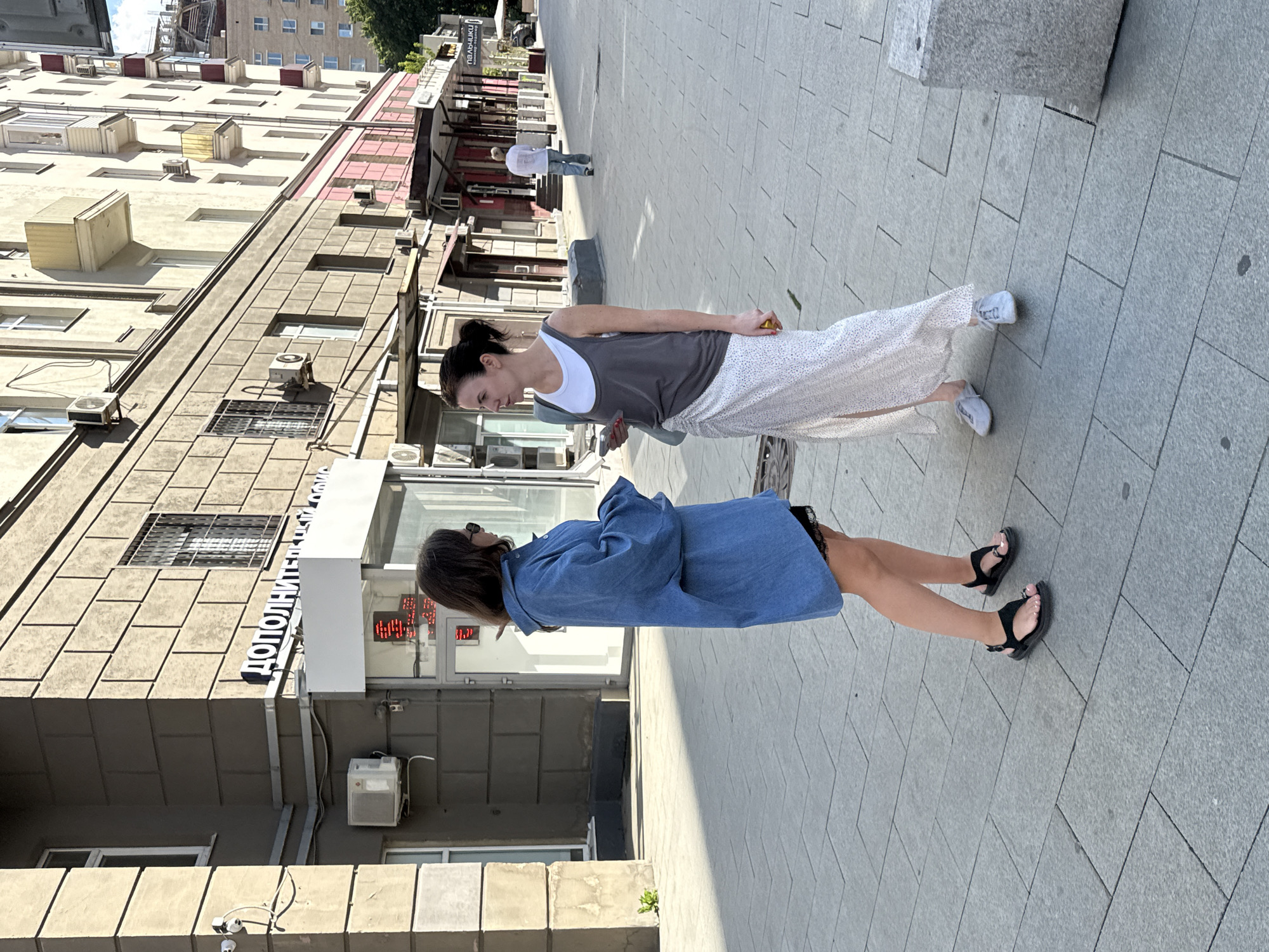 Two women are standing on a paved sidewalk in front of a building on a sunny day. The woman on the left is wearing a loose-fitting blue denim shirt dress and black sandals. She has long brown hair and is looking to her right, away from the camera. The woman on the right is wearing a grey tank top over a white t-shirt, a long white skirt with a slit, and light-colored sneakers. She has dark hair pulled back and is looking to her left, towards the other woman, holding a phone in her right hand.
The building behind them appears to be a commercial or office building with large windows and signage in Cyrillic. The architecture suggests an urban setting, likely in Moscow, Russia, given the presence of Cyrillic script. The sidewalk is made of large, grey rectangular paving stones. In the background, other people can be seen walking, and there are more buildings and some trees visible. The lighting indicates it is daytime, with strong sunlight casting shadows.