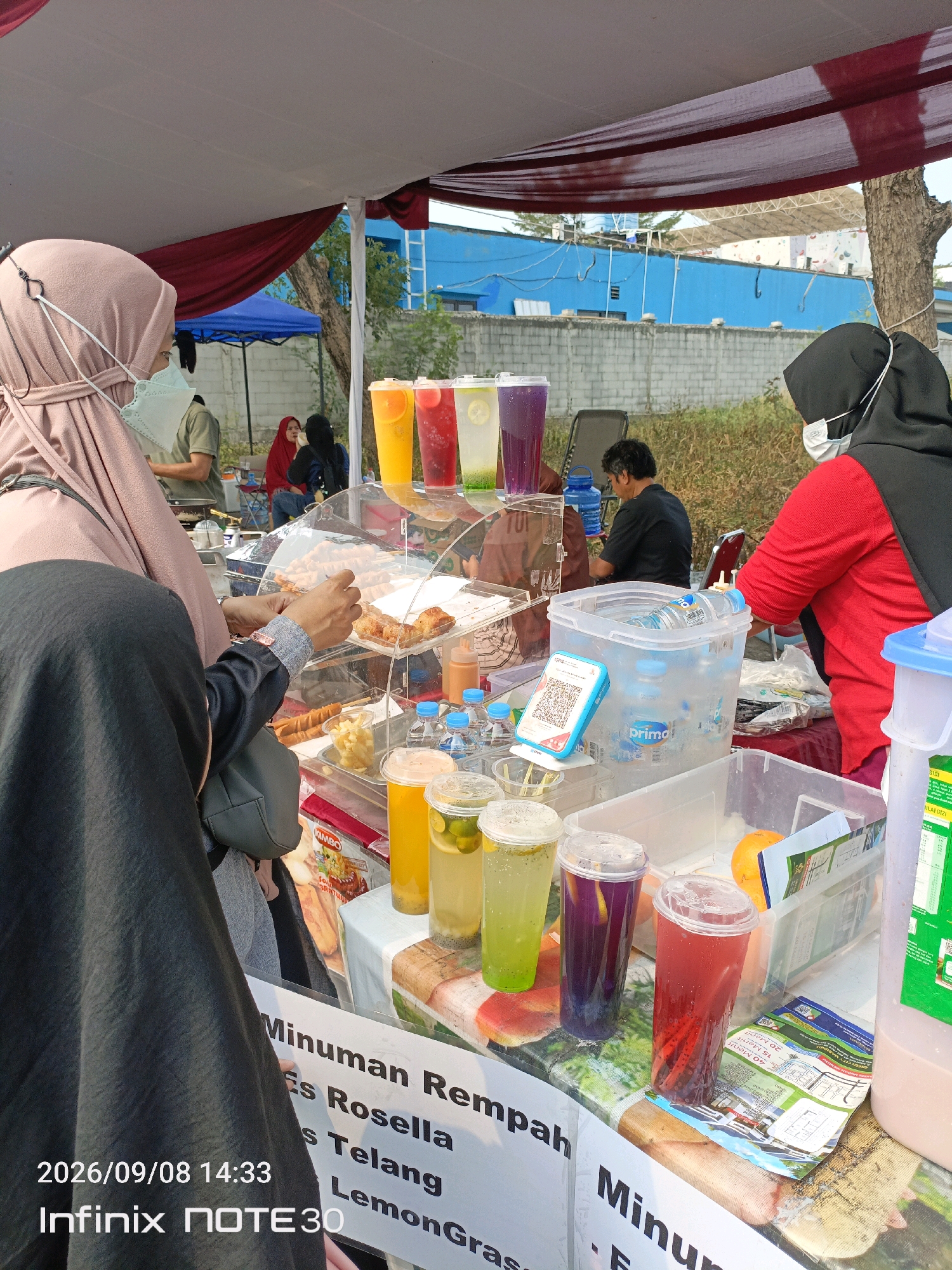 A group of people are at an outdoor food stall in East Jakarta, Indonesia. The stall is covered by a canopy and displays various drinks and food items. In the foreground, a woman in a black hijab and a red shirt, wearing a face mask, stands behind the counter, interacting with customers. To her left, another woman, also wearing a hijab and face mask, is handing over payment. Several colorful drinks in plastic cups are visible on display, along with skewers of food. In the background, other individuals are seen browsing the stall, and a blue building is visible behind them. The date "2026/09/08 14:33" and "Infinix NOTE30" are overlaid on the image, suggesting it may be a phone-captured photo with metadata.