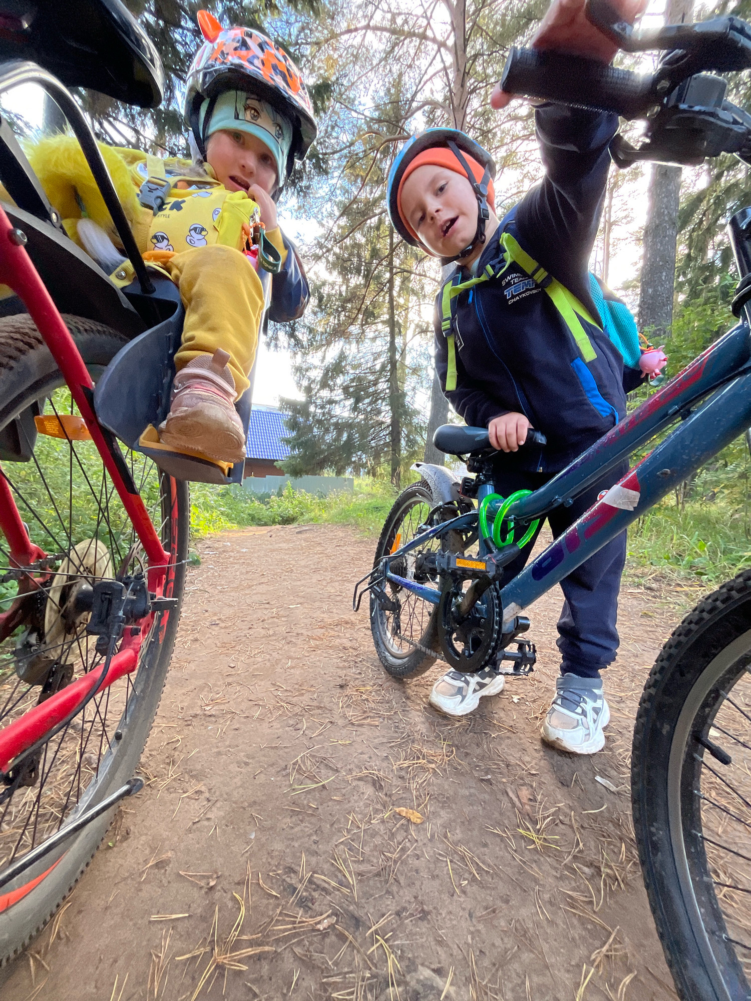 Two young children are on bicycles in a wooded area. The child on the left, a toddler wearing a yellow jacket and yellow pants, is seated in a bike seat attached to a red bicycle. The child is wearing a helmet with orange and black markings and a light blue bandana with an anime character on it. The child on the right, an older child, is standing next to a blue bicycle, holding the handlebars. This child is wearing a blue jacket, dark pants, and a helmet with an orange top. Both children are wearing athletic shoes. The bicycles are on a dirt path, surrounded by trees and dry pine needles. In the background, a residential area with a blue-roofed house and a fence can be seen. The image has been taken from a low angle, looking up at the children and bicycles. The location is identified as the city of Chaikovsky, Russia, based on text visible on the backpack of the child on the right.