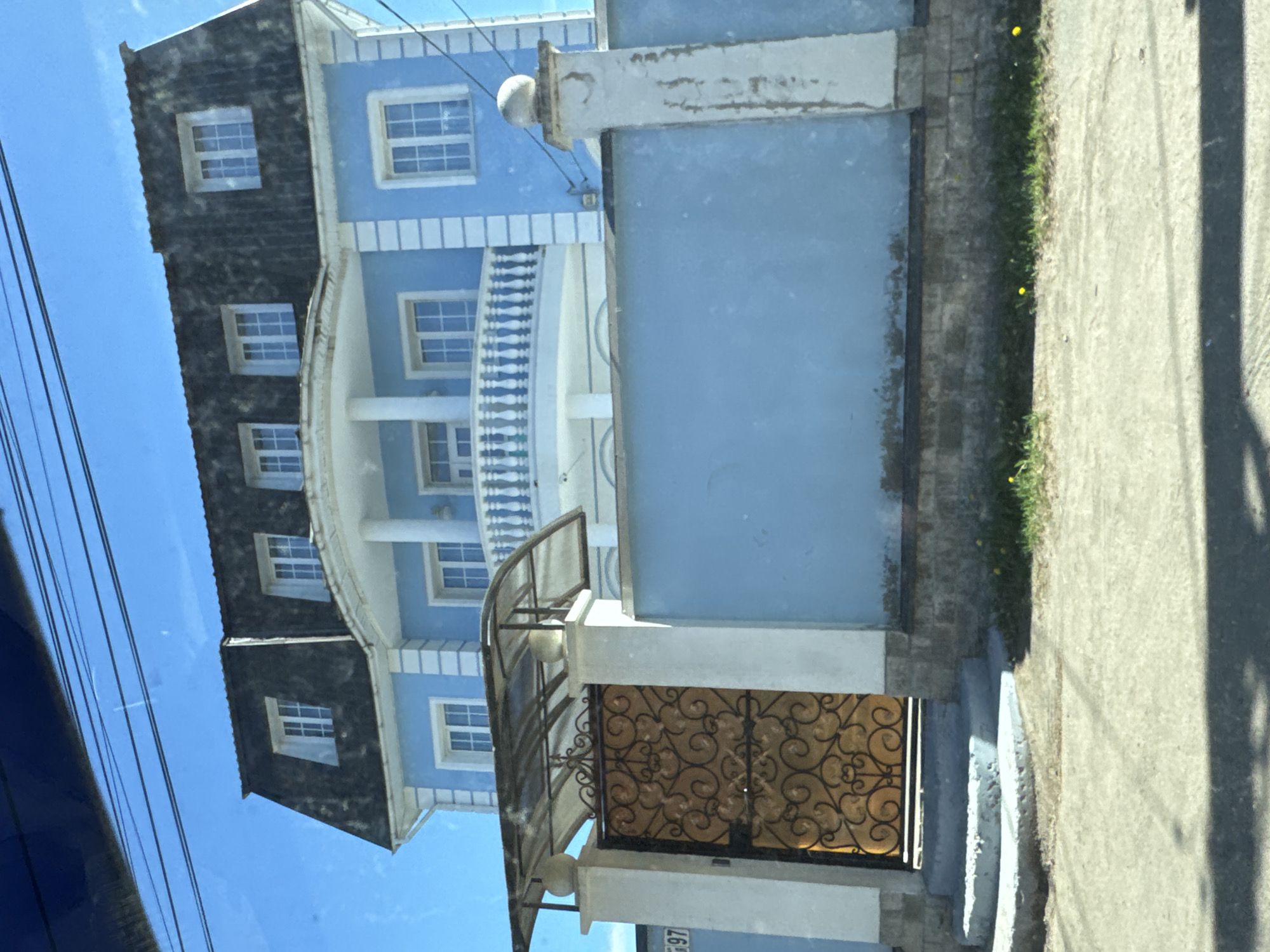 An exterior daytime shot in Pesochny, Russia, captures a two-story light blue house with a dark gray mansard roof and white architectural trim. The house features numerous rectangular windows with white frames and grids on both main floors and within the dormers of the roof. A prominent curved balcony with white balustrades and supporting columns is centered on the second floor. In the foreground, a light blue solid wall fence with white decorative pillars runs parallel to the street. An ornate, golden-colored wrought-iron gate with intricate scrollwork is integrated into the fence on the left, beneath a curved, translucent canopy. The house number "97" is visible on a white plaque attached to the left gate pillar. The ground level includes a concrete sidewalk or driveway in the immediate foreground, with a strip of grass containing small yellow dandelions bordering the fence. The sky overhead is clear and bright blue, traversed by multiple dark power lines. No individuals or discernible actions are present within the scene.