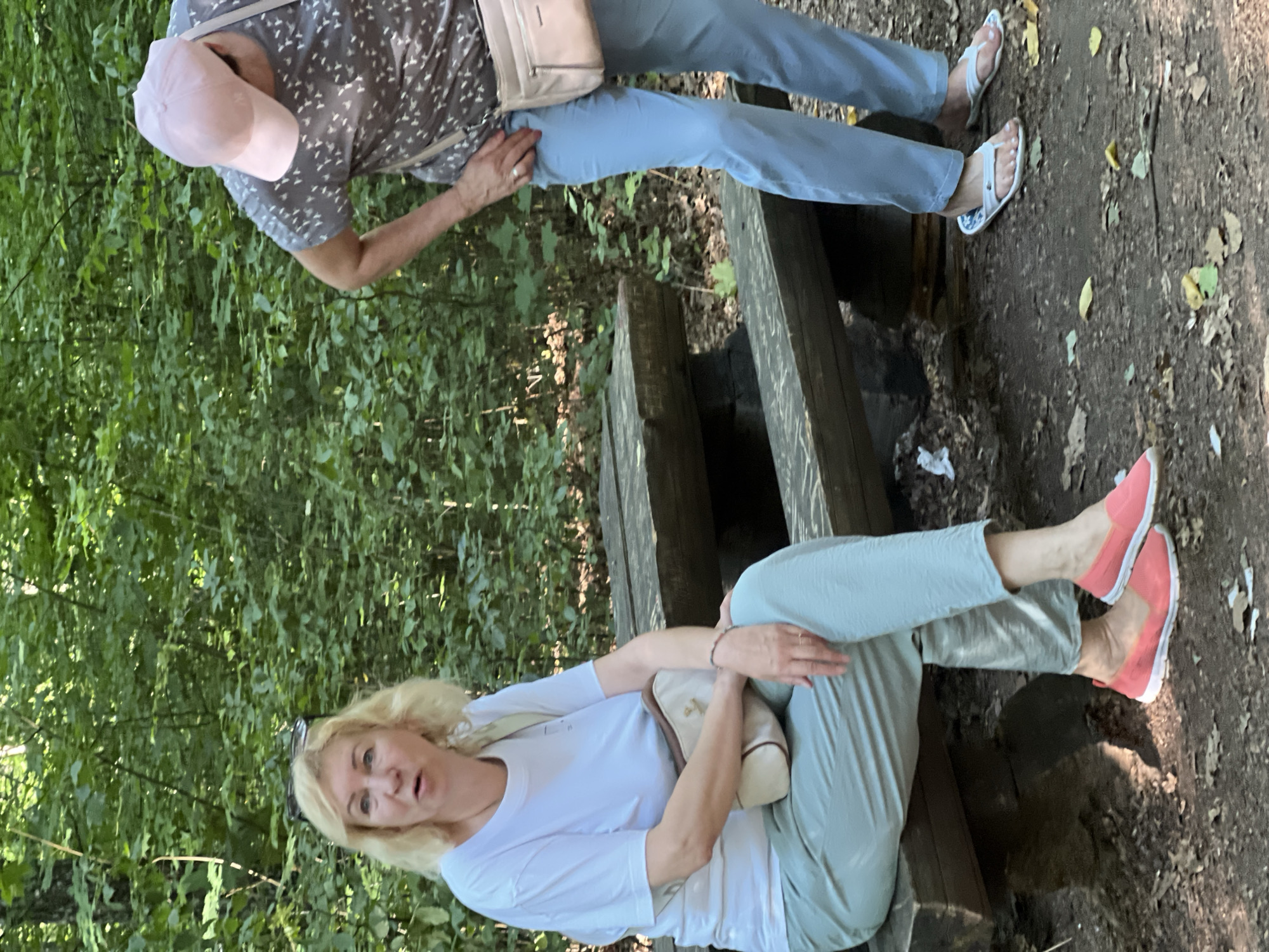 In the city of Сонячне, Ukraine, two women are present in a wooded area. One woman, with blonde hair, is seated on a wooden bench, wearing a white t-shirt, light green pants, and coral-colored slip-on shoes. She has a light-colored crossbody bag. The second woman, wearing a pink cap, a gray patterned t-shirt, and light blue pants, is standing to the right of the bench, bending slightly. She is also wearing a light-colored crossbody bag and flip-flops. The environment is a forest floor with dry leaves and dirt, and dense green foliage forms the background. A wooden picnic table is partially visible behind the seated woman.