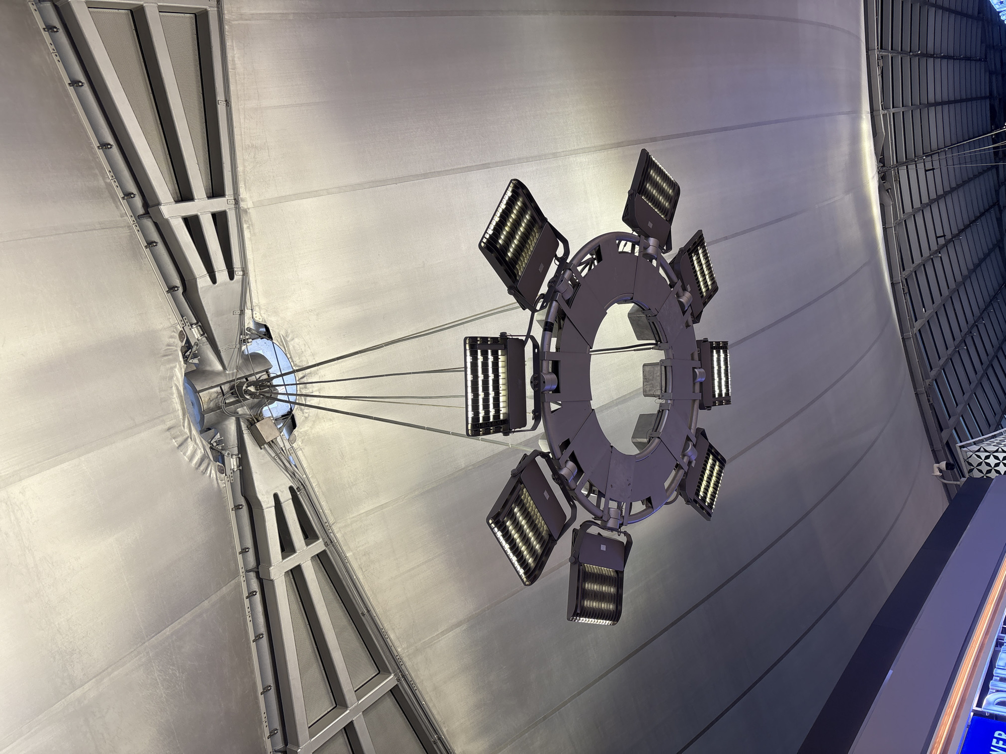 A circular, modern chandelier with multiple LED lights hangs from a large, white, tent-like ceiling. The chandelier is made of a gray metallic material and is suspended by thin wires. The ceiling is constructed of fabric stretched over a metal frame. Below the chandelier, on the right side of the image, is a glimpse of a dark, patterned structure and a security camera. The bottom left corner shows a brightly lit blue and white sign.