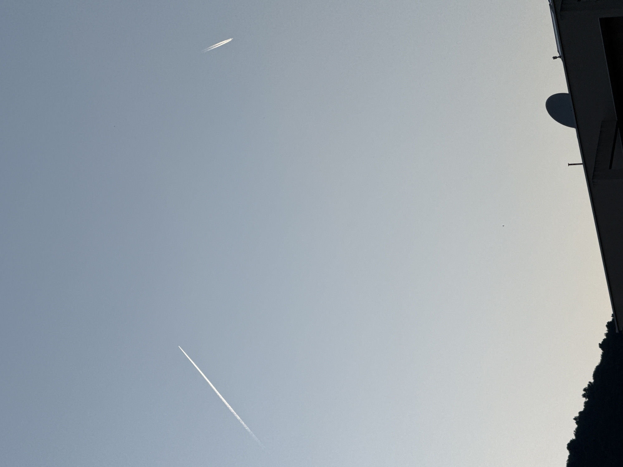 The image captures a clear sky above the city of Budva, Montenegro. Two contrails from aircraft are visible, one extending diagonally across the upper left portion of the frame and the other further to the right. The lower section of the image shows the silhouetted rooftops of buildings, with a prominent satellite dish on one of them. A dark, tree-covered hillside is also visible on the left. The scene is characterized by soft, natural light, suggesting either early morning or late afternoon. There are no people or animals present in the frame.