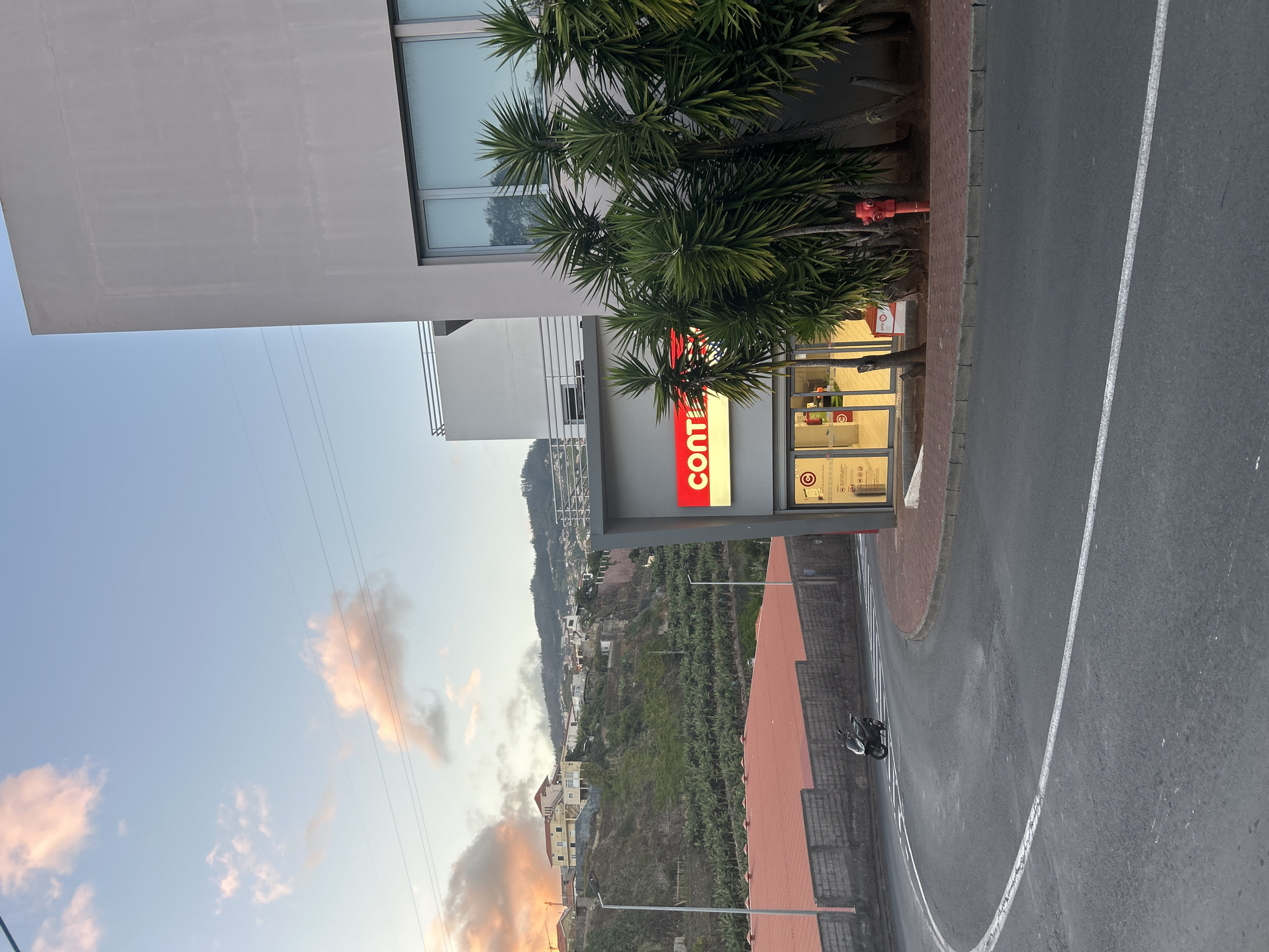 The image shows a view of a Continente Bom Dia supermarket in Câmara de Lobos, Portugal. The supermarket is located on a hill slope. It features a red "Continente" sign and a glass-fronted store. The building is a modern, multi-story structure. The surrounding environment includes trees next to the supermarket and a sloped hillside with buildings and trees extending into the distance. A road runs alongside the supermarket, with white lines marking lanes. A moped is parked nearby. The sky in the background shows sunset hues.