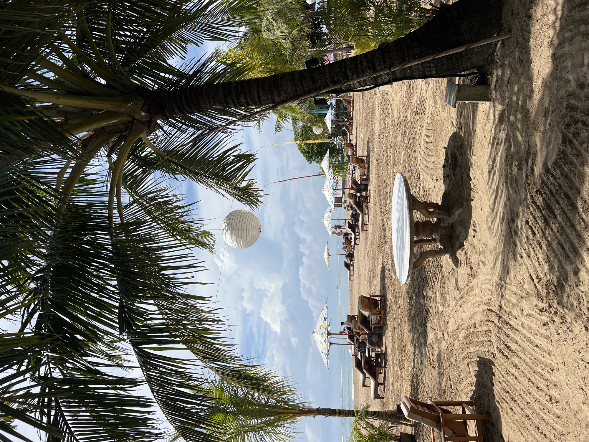 This media file captures a vibrant tropical beach scene, viewed from beneath the canopy of lush palm trees. The image is oriented to show a wide expanse of sandy beach extending towards a calm ocean under a bright blue sky with scattered white clouds.
In the immediate foreground, dark green palm fronds and thick palm trunks frame the left, top, and bottom of the composition, creating a natural archway. These palms cast distinct shadows on the sandy ground, which is textured with ripples and imprints.
The mid-ground is dominated by the beach, where several people are seen relaxing on wooden lounge chairs and benches. These are accompanied by various beach umbrellas, some closed and some open, providing shade. On the right side of the frame, a distinct round white table with an ornate carved base stands on the sand. A large white paper lantern hangs from a line in the upper left section of the sky.
In the background, the serene, light blue ocean meets the horizon. The sky above is clear and bright. The scene suggests a leisurely day at a beach resort, with people enjoying the sun and calm waters. The sunlight indicates it is daytime, likely late morning or early afternoon. No visible text is present in the image.