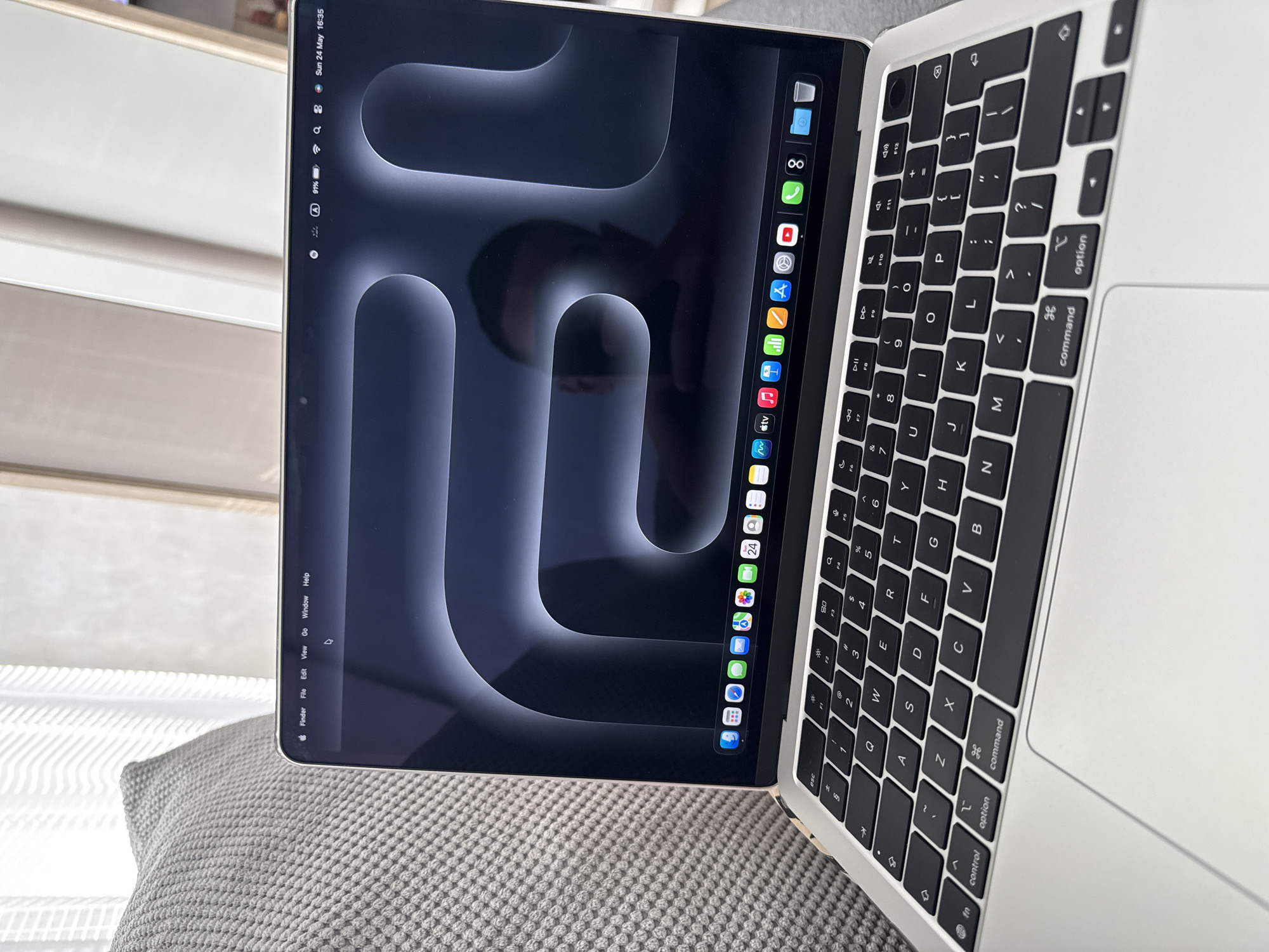 The image shows a modern Apple MacBook laptop resting on a textured gray surface, likely a cushion or sofa. The laptop screen displays a dark-themed wallpaper with rounded, abstract shapes, and a dock of applications is visible on the left edge. The screen also shows system information like battery percentage (91%) and the date "Sun 24 May 16:35." The keyboard, which is black with white lettering, is partially visible, along with the trackpad. The laptop is the central focus of the image, with only a blurry suggestion of the location.