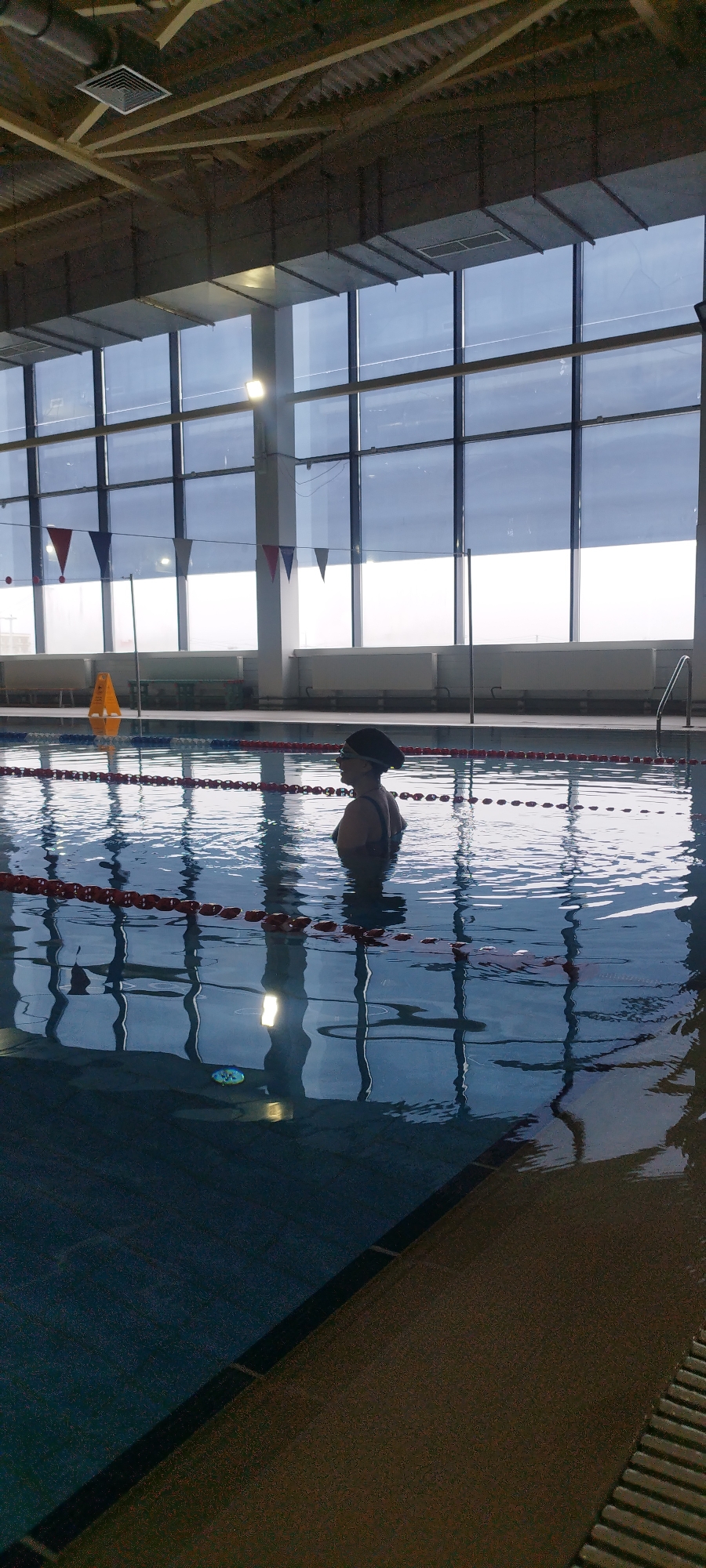 The image depicts an indoor swimming pool, likely located in Yakutsk, Russia, based on the architectural style common in colder regions and the presence of indoor facilities necessitated by the climate. The pool is divided into lanes by red-and-white floating dividers. A woman is standing in the water, facing left, wearing a dark swimming cap and goggles. She appears to be stationary. Large windows line the back of the pool area, showing a muted daylight scene outside. The pool water is reflecting the lights and the window frames. The foreground shows the edge of the pool with a dark brown textured surface. A yellow warning cone is visible on the side of the pool in the background.