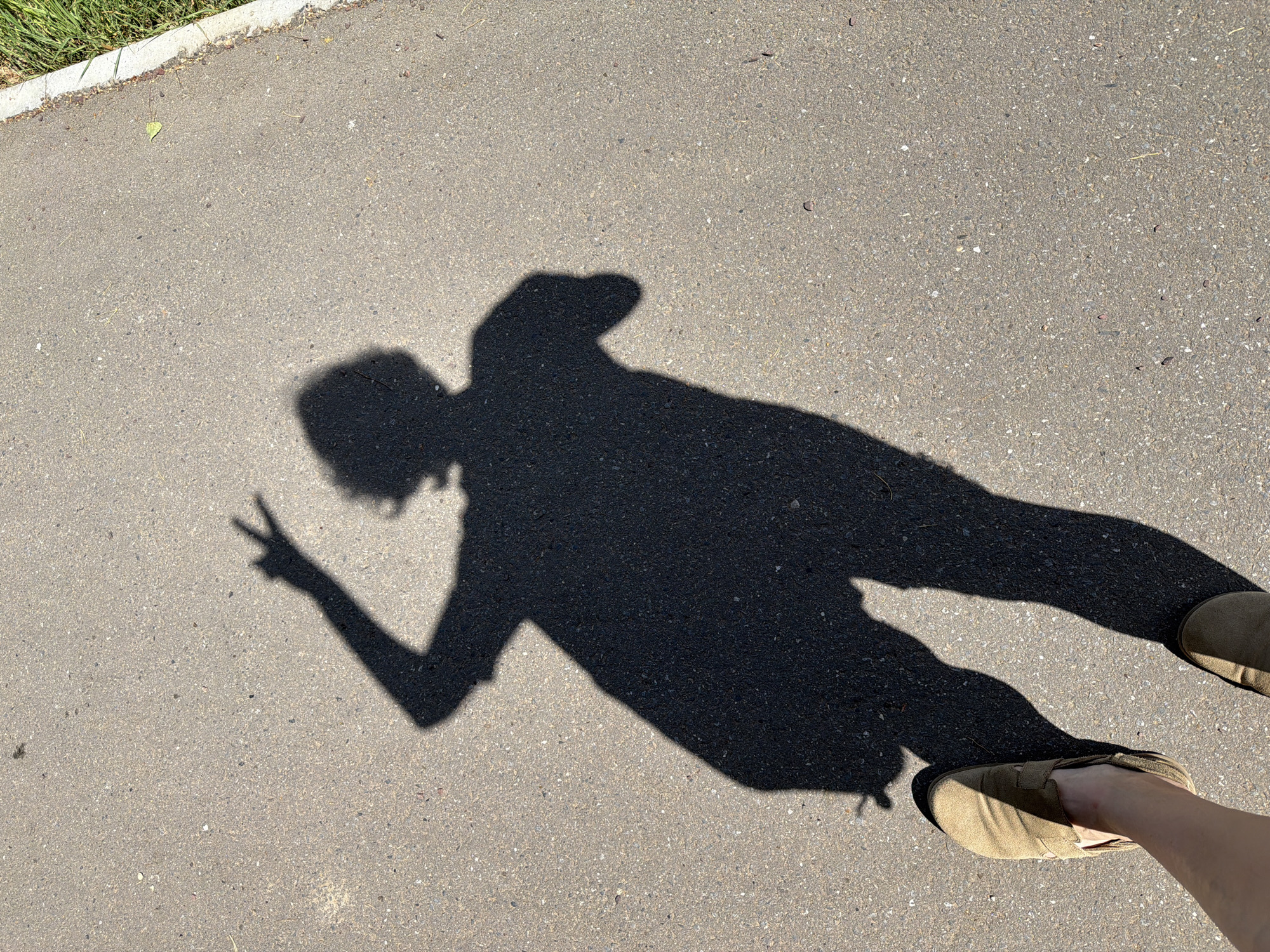 The image shows the shadow of a person making a peace sign, with a foot visible in the lower right corner wearing a tan shoe. The shadow and foot are on an asphalt surface. There is grass visible in the upper left corner. The image location is Chaikovsky, Russia.