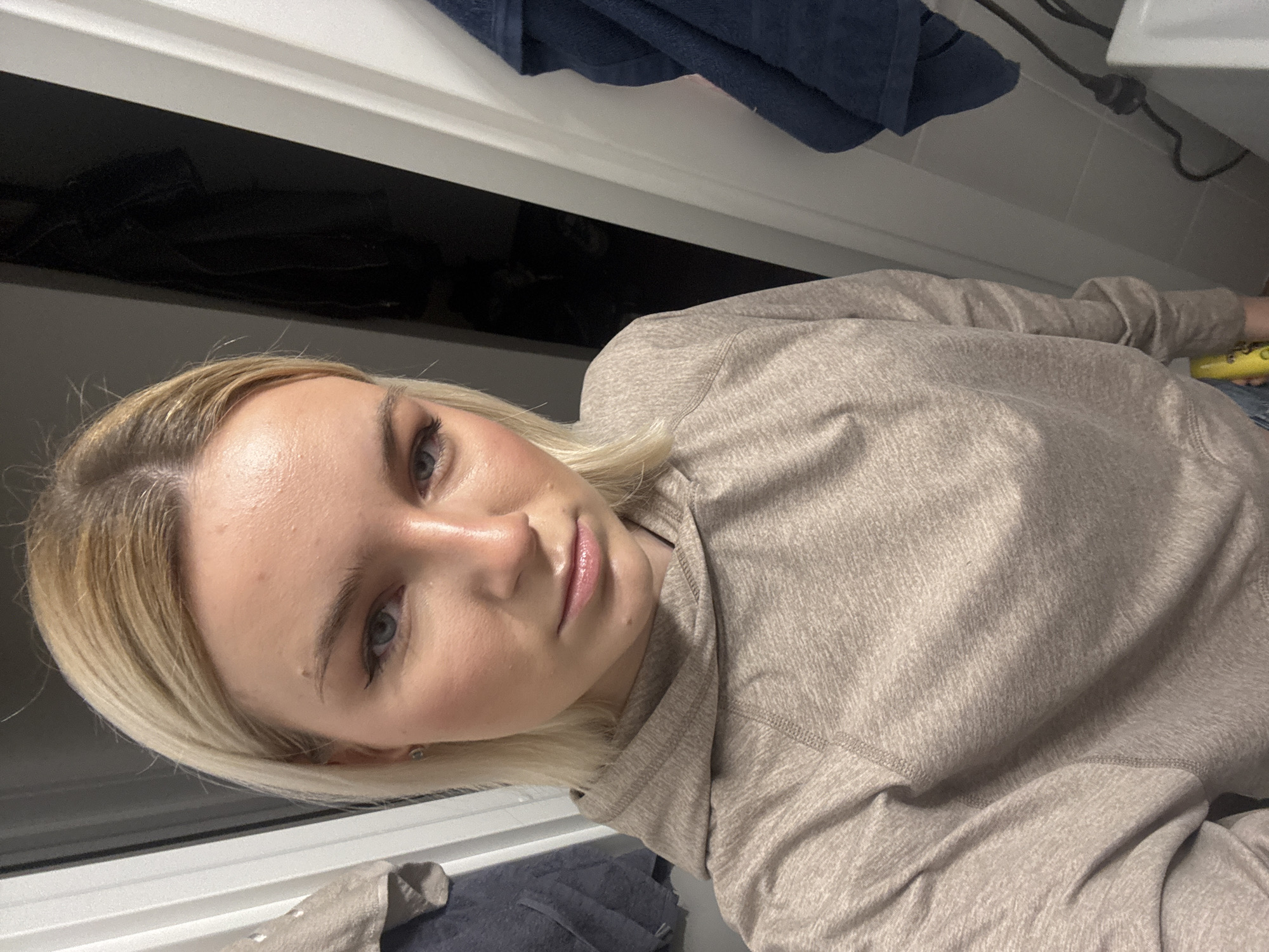 A young woman with fair skin and blonde hair is captured in a close-up, seemingly in a bathroom setting. She wears a light brown, hooded sweatshirt. Her gaze is directed towards the camera. The background reveals a white bathroom cabinet, a dark blue towel hanging above, and a power cord running along the upper right. A window is visible behind her head, with what appears to be a blue/gray cloth near the bottom left. The city of Unknown, Russia, is not determinable from the image.