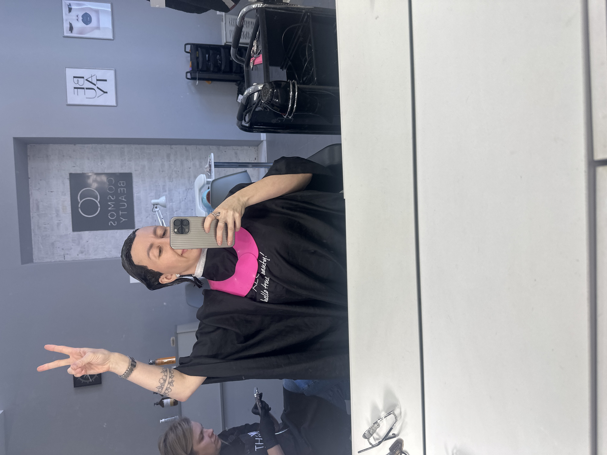 A mirror selfie captures a person in a hair salon in Votkinsk, Russia. The image is taken from the perspective of the person in the salon chair, reflecting a large mirror that dominates the right side of the frame.
The main subject is a woman, reflected in the mirror, taking the selfie with a gray smartphone held in her left hand. Her hair appears dark and wet, suggesting a hair treatment is in progress. She is wearing a black cape or smock over a light-colored top, and a pink neck collar is visible around her neck. Her right hand is raised, forming a peace sign, and a tattoo resembling a floral band is visible on her right forearm. She has a slight smile, indicating a positive mood.
In the background reflection, a black hair salon chair and a black salon cart with various products and tools can be seen. The walls are grey, adorned with two framed pictures. One picture features abstract facial art, while the other displays mirrored text that reads "BE YOU." Below these, another mirrored sign on a brick-textured wall section shows "BEAUTY COSMOS" with an interlocking circle logo. A round black clock is visible on the wall to the left of the main subject.
Partially visible in the lower left reflection, another person, likely a stylist, is seen wearing black gloves, suggesting an ongoing service. A pair of eyeglasses rests on a surface in the bottom right of the reflection. The black cape worn by the main subject has mirrored white text that reads "hello true party!". The overall setting is a modern and functional hair salon. The lighting is bright, suggesting it is daytime.