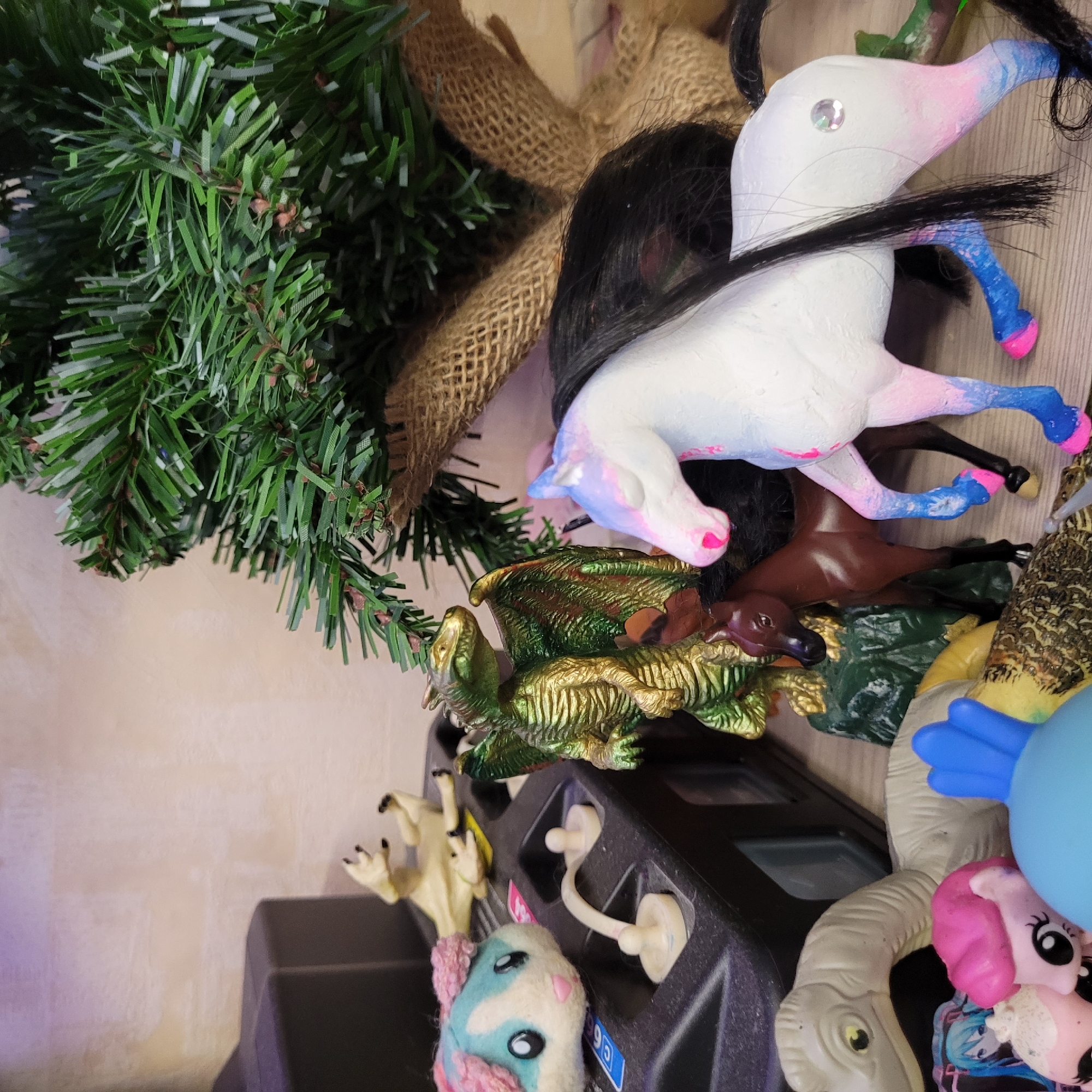 An image captures a close-up, top-down view of a collection of toys and artificial greenery, rotated 90 degrees clockwise.
On the left, a vibrant green artificial Christmas tree branch with small brown pine cones is visible, rooted in a light brown burlap base. To its right, a white unicorn toy with a black mane and tail lies on its back. The unicorn's hooves and muzzle are painted in gradient shades of pink and blue, and a clear, iridescent gem is affixed to its forehead.
Below the unicorn, a brown horse or pony toy is partially visible, lying on its side. Next to it, a detailed green and gold dragon toy with prominent scales and wings is positioned, seemingly resting on a dark green base.
Further down and to the right, a black plastic object with multiple square openings and white hooks is present, possibly a toy organizer or a power strip. A small, light blue and white plush animal with pink ears and black eyes is nestled against this object. Above it, a light-colored toy with visible claws is seen.
On the far right and bottom, various other toys are clustered: a gray dinosaur-like figure with a visible head and neck, a pink pony toy (resembling My Little Pony), a bright blue round object, and a small figure with blue hair. A blue sticker on the black plastic object bears the text "C5".
The background behind the objects is a plain, light-colored surface, possibly a wall or tabletop. The overall impression is a vibrant and somewhat cluttered arrangement of children's playthings.