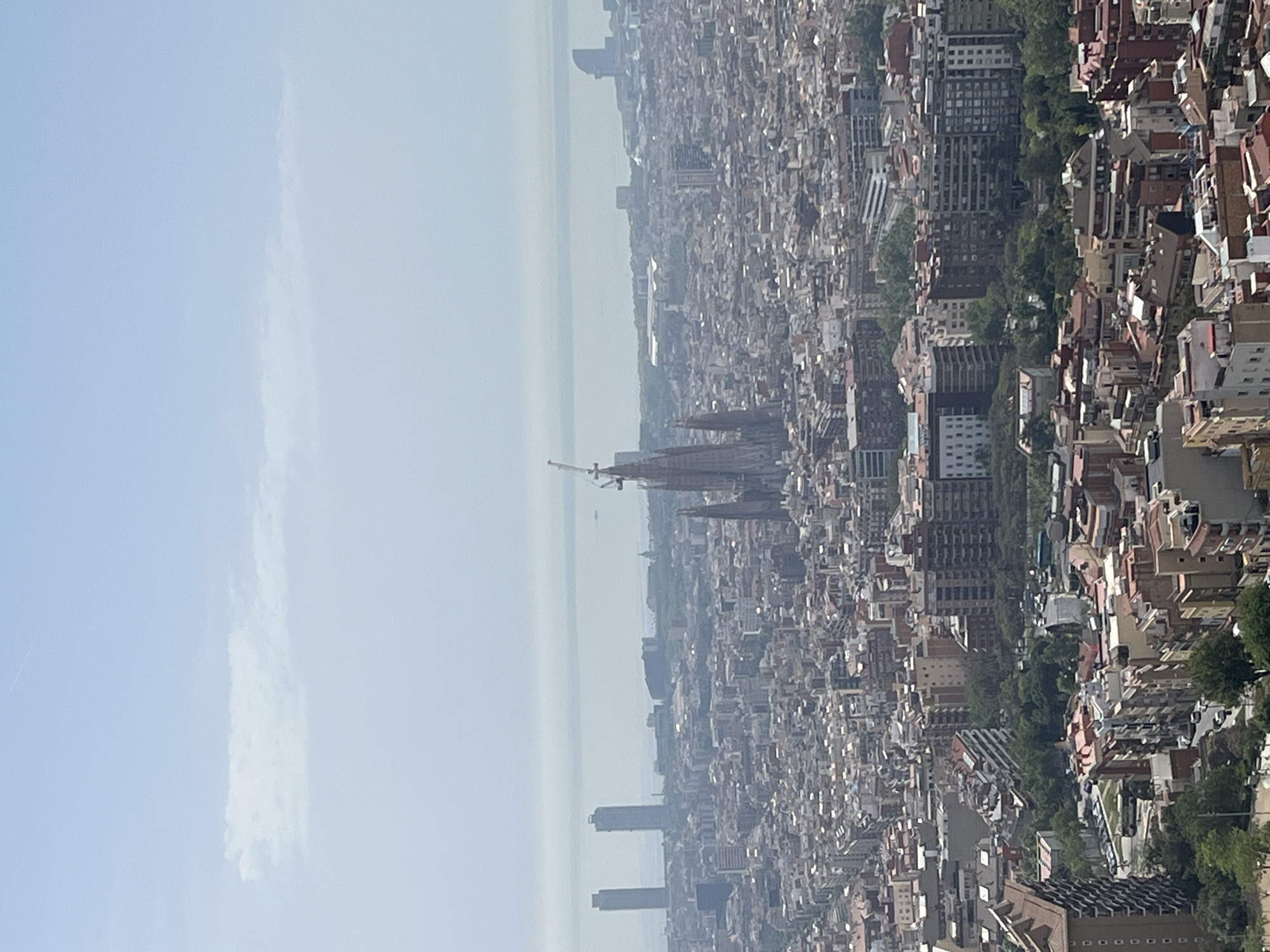 The image presents a panoramic aerial view of a sprawling cityscape, identified as Barcelona, Spain, taken from a high vantage point. The perspective looks over a vast expanse of buildings extending towards a hazy horizon, which appears to be the Mediterranean Sea.
The urban landscape is densely packed with a multitude of buildings, characterized by varied architectural styles and numerous reddish-brown tiled roofs. Prominently positioned in the mid-ground is the Sagrada Familia cathedral, its iconic spires and a visible construction crane making it an unmistakable landmark. Further towards the horizon, several taller, more modern high-rise buildings punctuate the skyline. Interspersed within the built-up area are patches of green vegetation, indicating parks or tree-lined avenues.
The sky above is a pale, diffused blue, largely clear but with subtle, wispy white clouds. The overall atmospheric condition is hazy, reducing visibility, particularly towards the distant horizon where the sea meets the sky. This haze creates a soft, diffused light over the city.
The scene depicts a quiet, expansive view of the city during daytime, likely under bright but not directly sunny conditions due to the atmospheric haze. No specific human activities or individuals are discernible given the vast distance of the viewpoint. There is no visible text present in the image.
